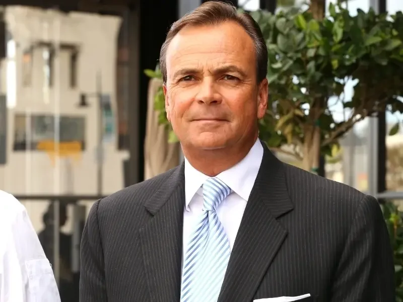 A middle-aged man with dark hair, wearing a dark pinstripe suit, white shirt, and light blue tie, standing outdoors near greenery and glass buildings.