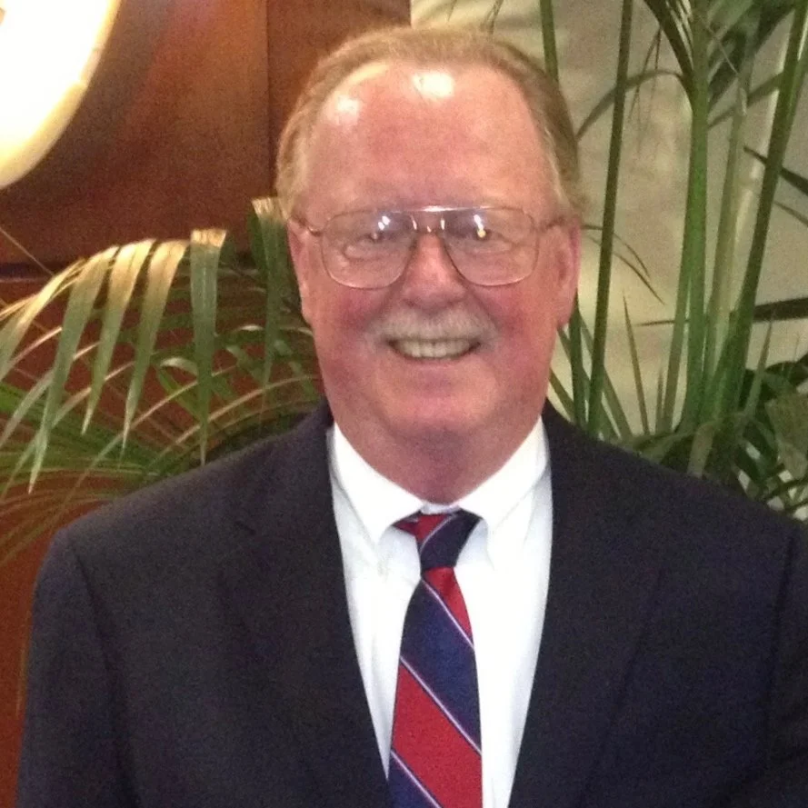 A smiling elderly man with glasses, wearing a dark suit, white shirt, and a diagonally striped red, blue, and white tie, standing in front of green plants.