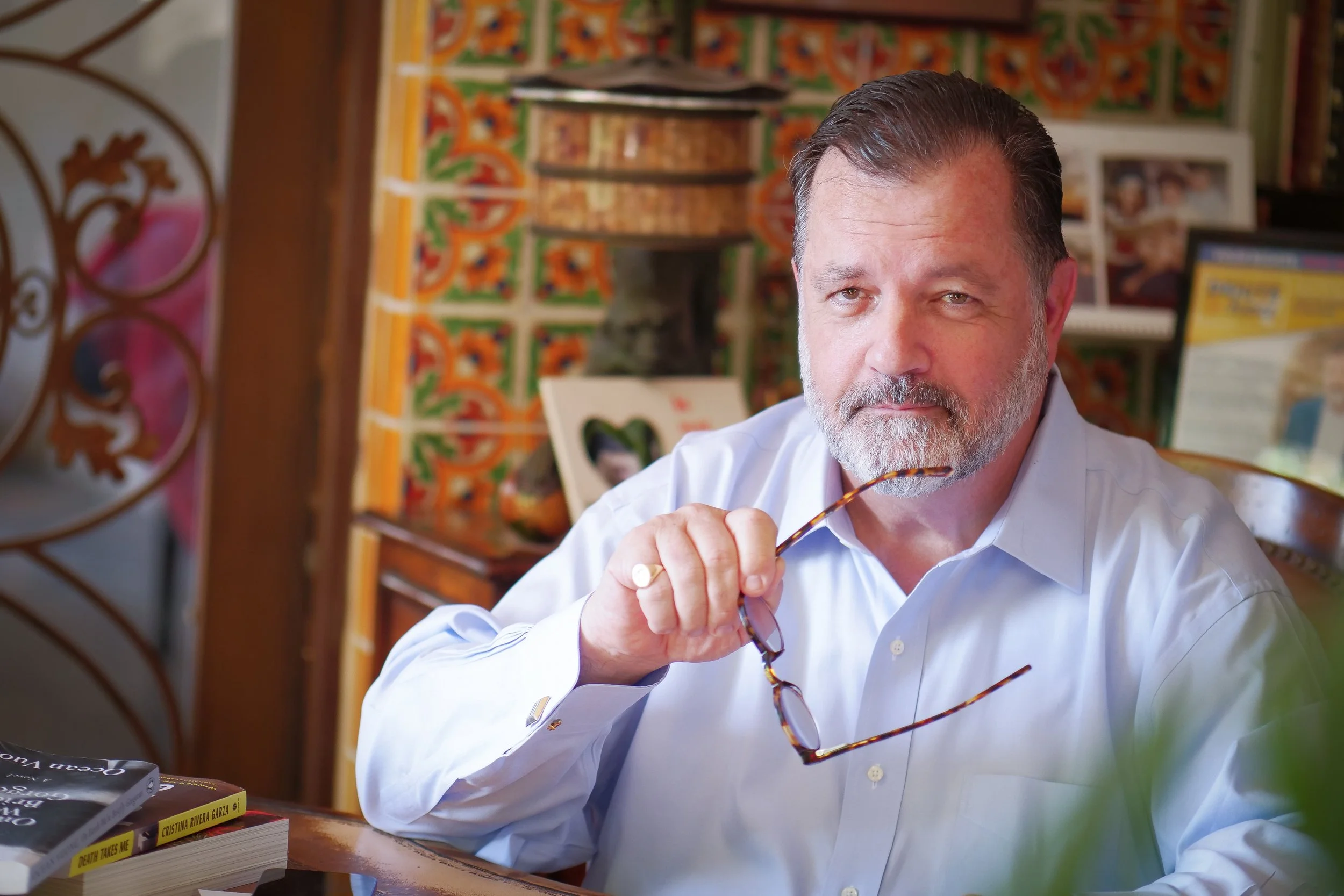 A middle-aged man with gray hair and a beard, wearing a white shirt, sits at a table in a colorful, decorated room, holding glasses in his right hand.