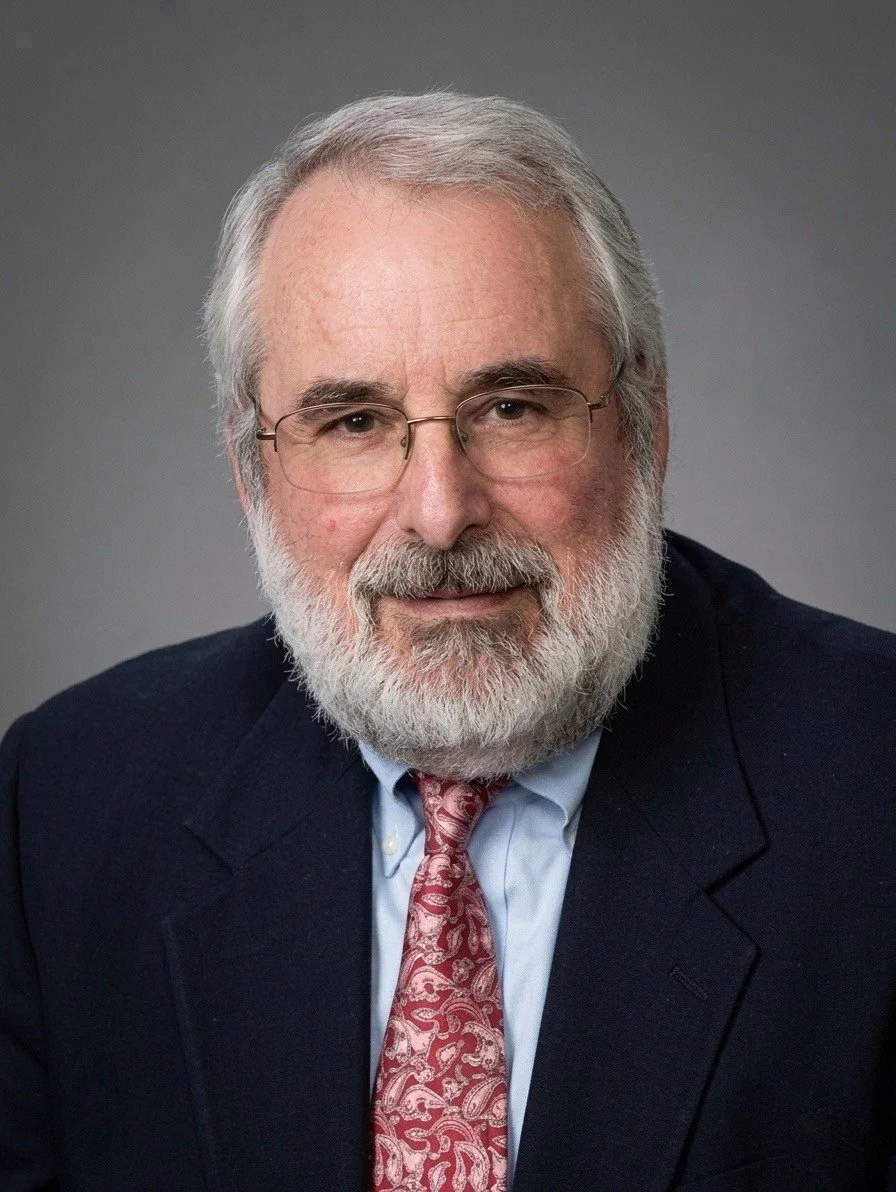 Portrait of an older man with gray hair and beard, wearing glasses, a dark suit, light blue shirt, and patterned red tie against a neutral background.