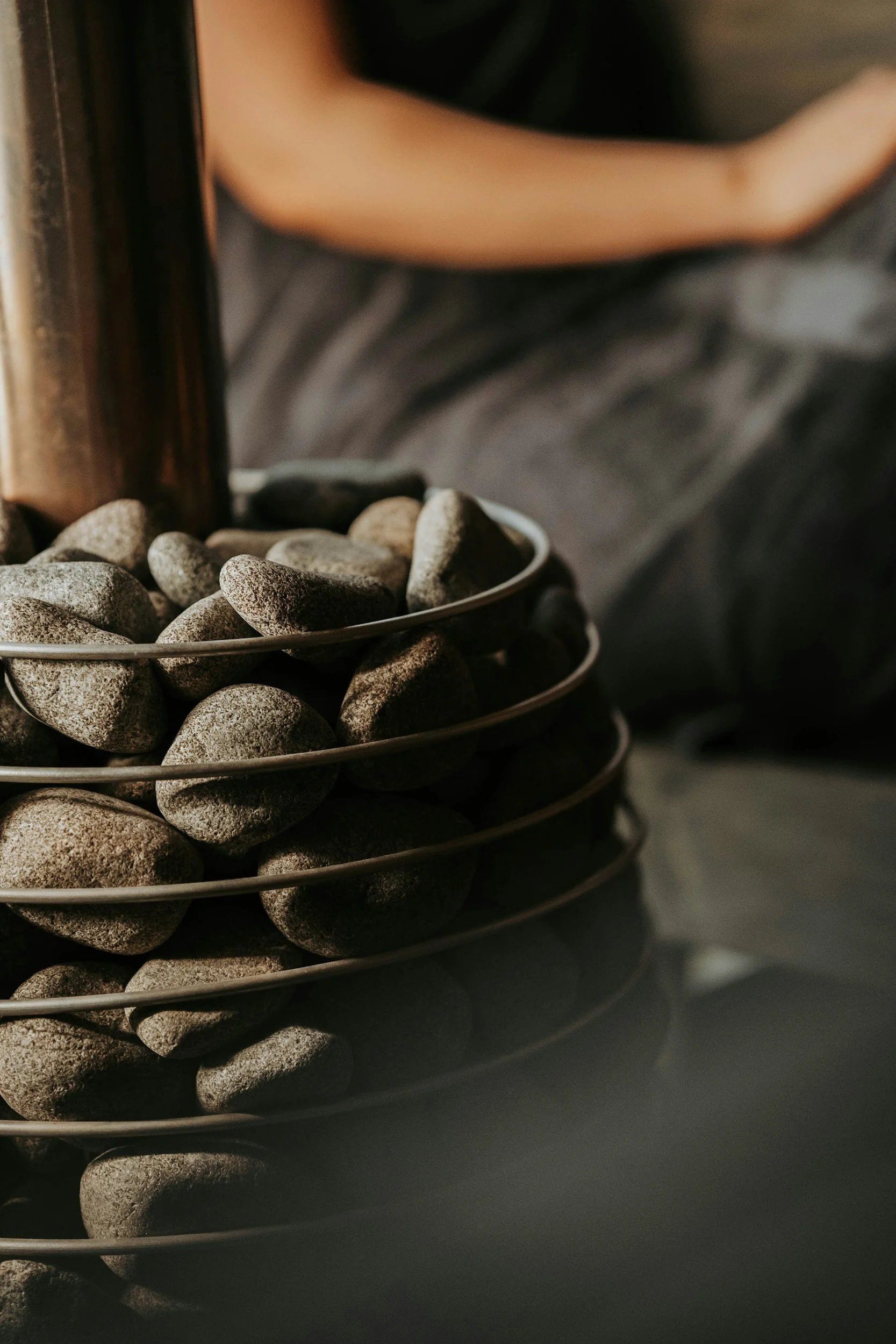 Close-up of a sauna heater filled with small, smooth gray stones, with part of a person in dark clothing and an arm visible in the background.
