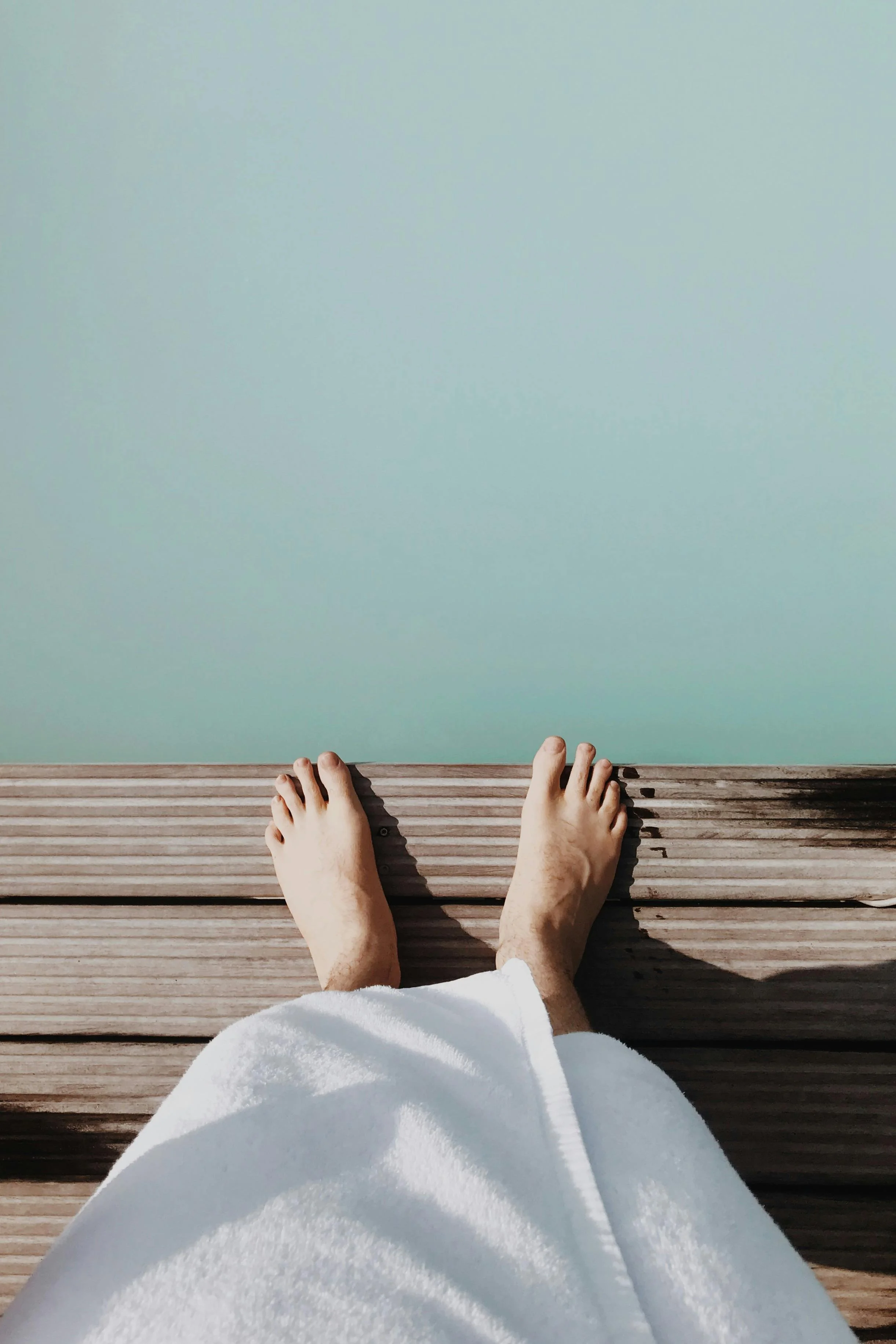 A person's bare feet and ankles visible, standing on a wooden deck with horizontal planks, near a light green wall, partially clothed in a white garment.