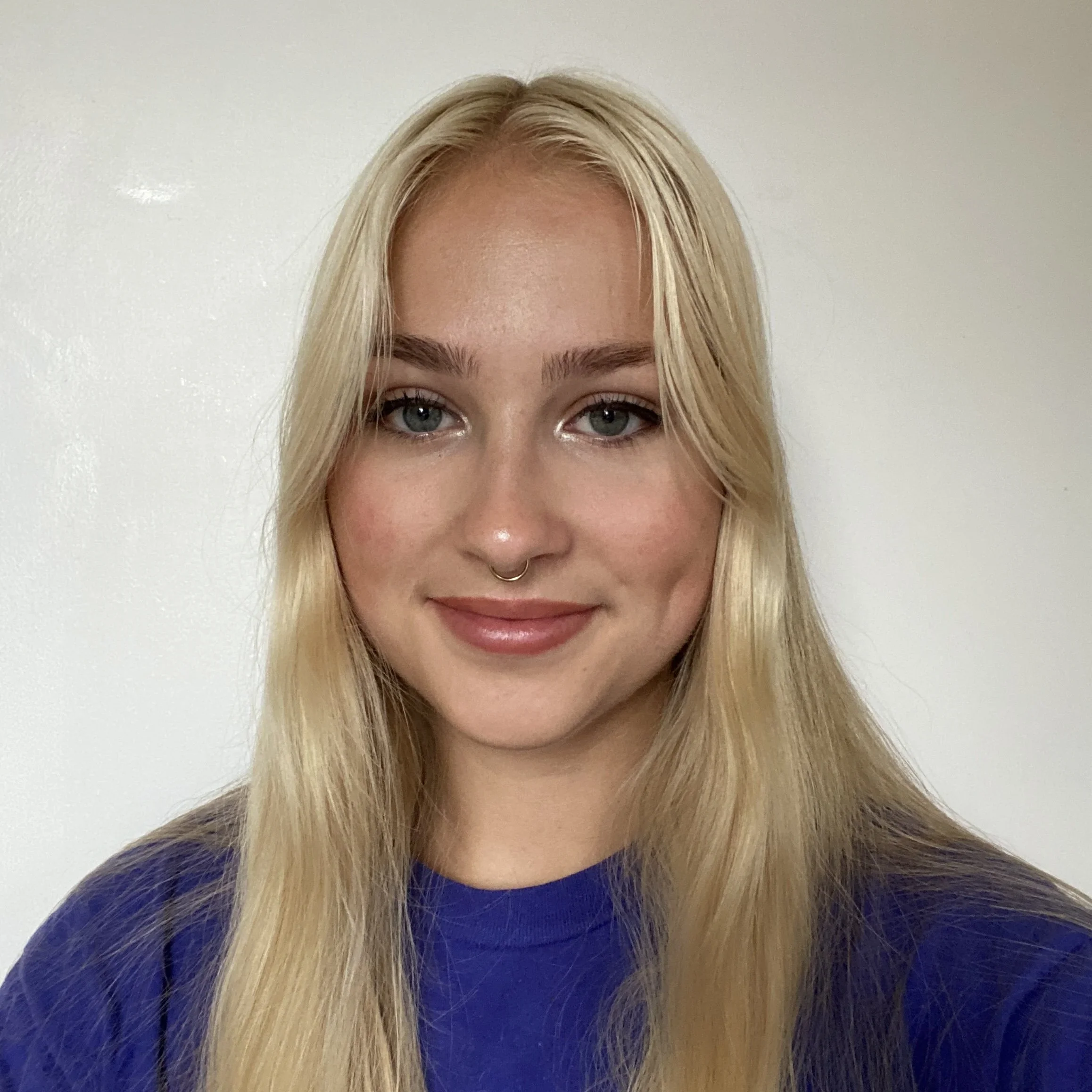 A woman with blonde hair, blue eyes, freckles, and a nose ring smiling and wearing a dark blue top against a white wall background.