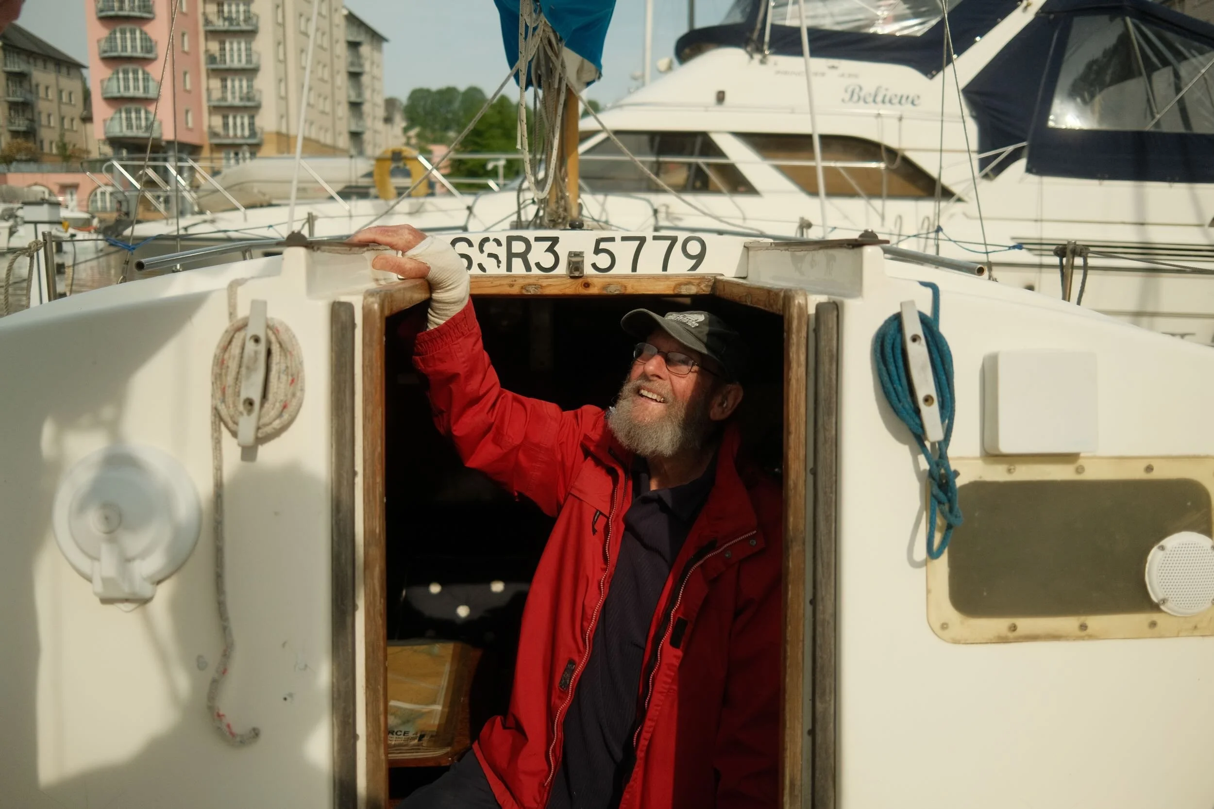 A man with glasses, a beard, and a cap, smiling while working on a boat, with buildings and other boats in the background.