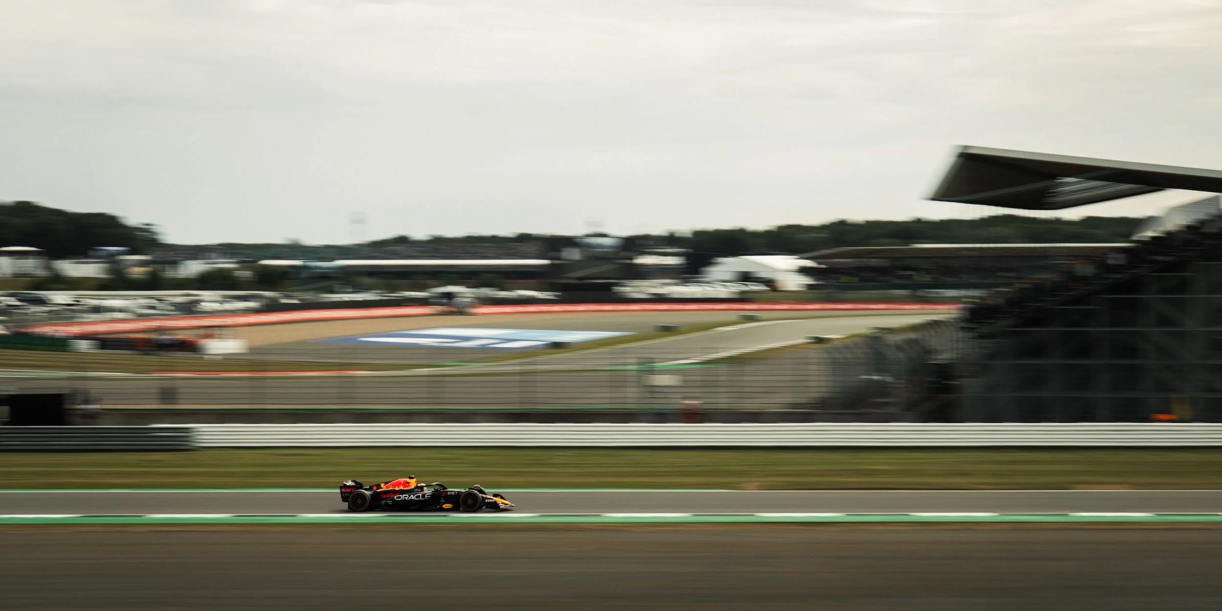 A racing car on a race track with a grandstand and control tower in the background, blurred to show speed.