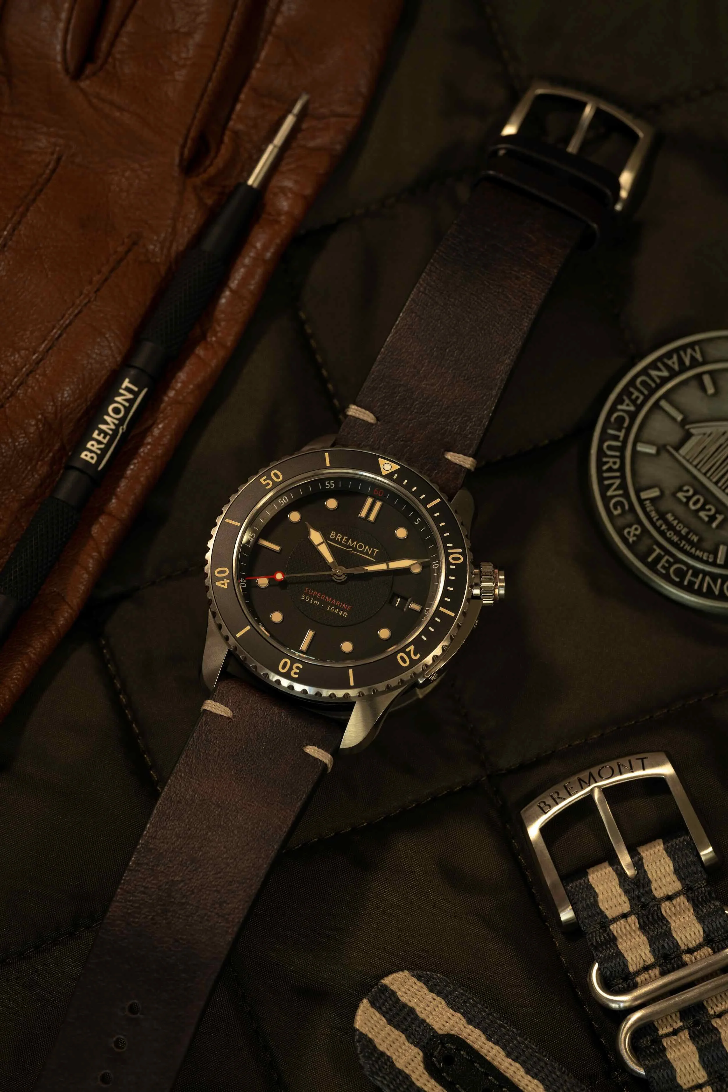 Close-up of a Bremont wristwatch with a dark strap, a brown leather belt, and other watch straps on a black surface.