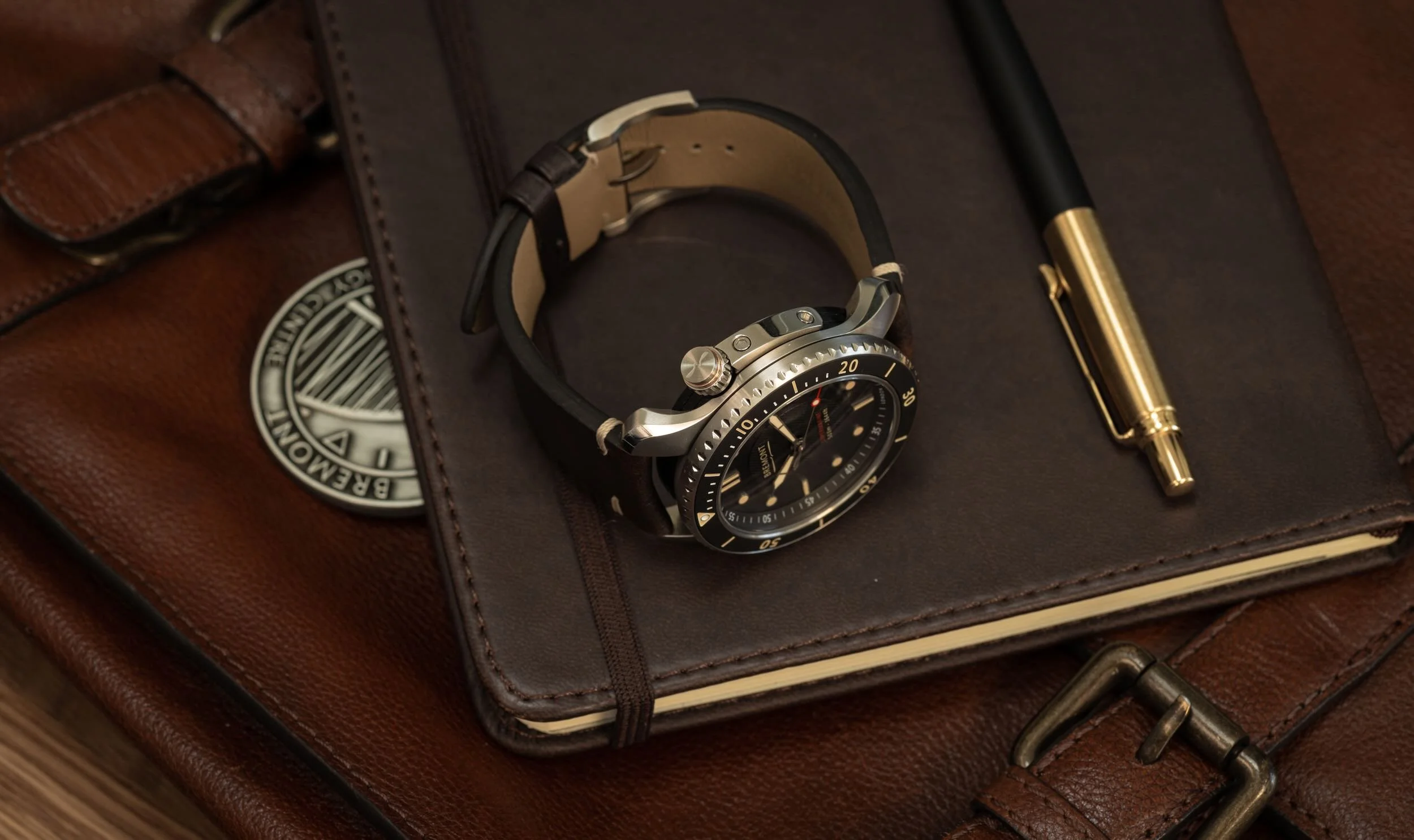 A wristwatch with a black and gold bezel, black strap, and multiple dials on the watch face, placed on a dark leather surface inside a brown leather carrying case with a partially visible logo.