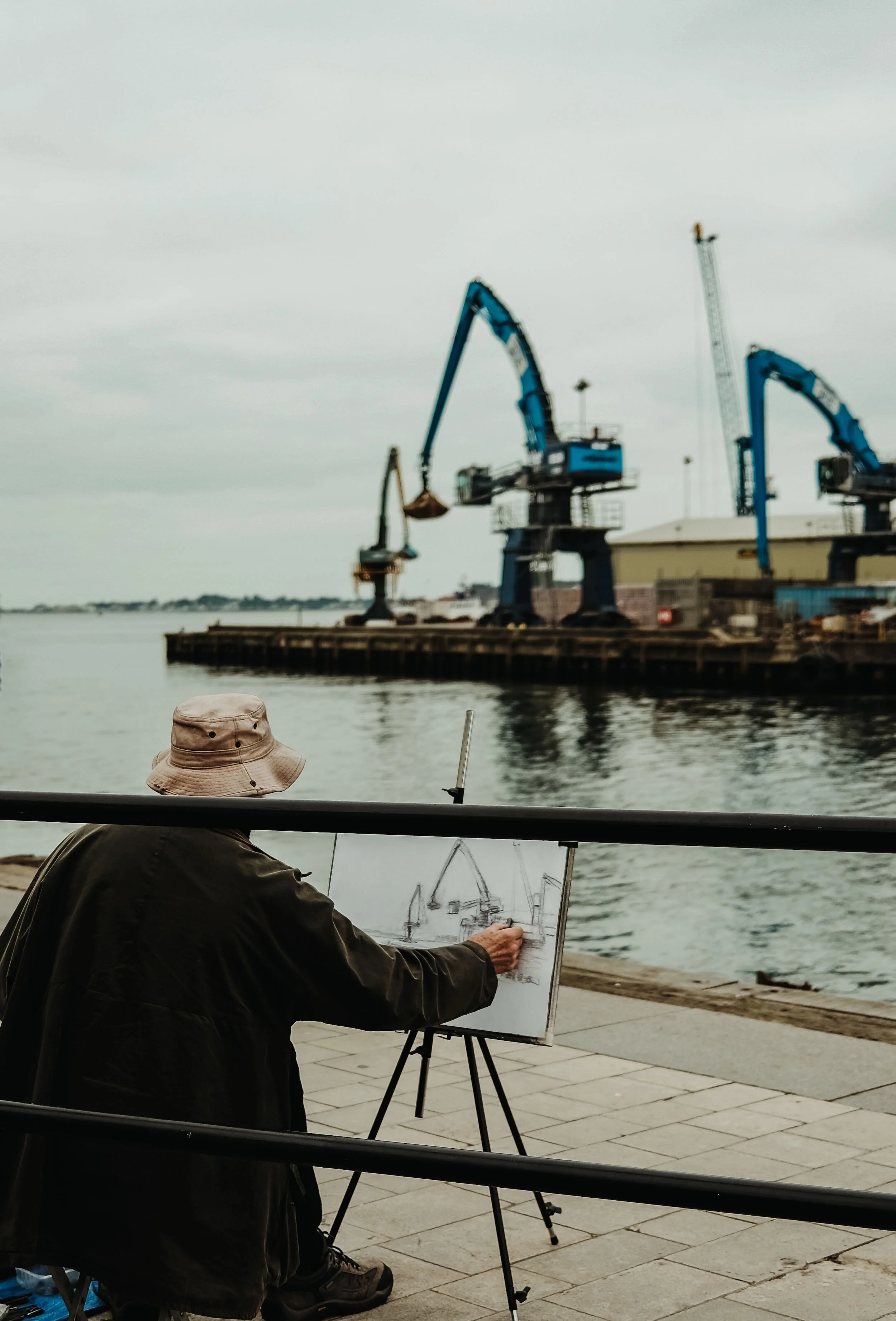 A man wearing a bucket hat and dark jacket painting a waterfront scene on an easel, with water and industrial cranes in the background.
