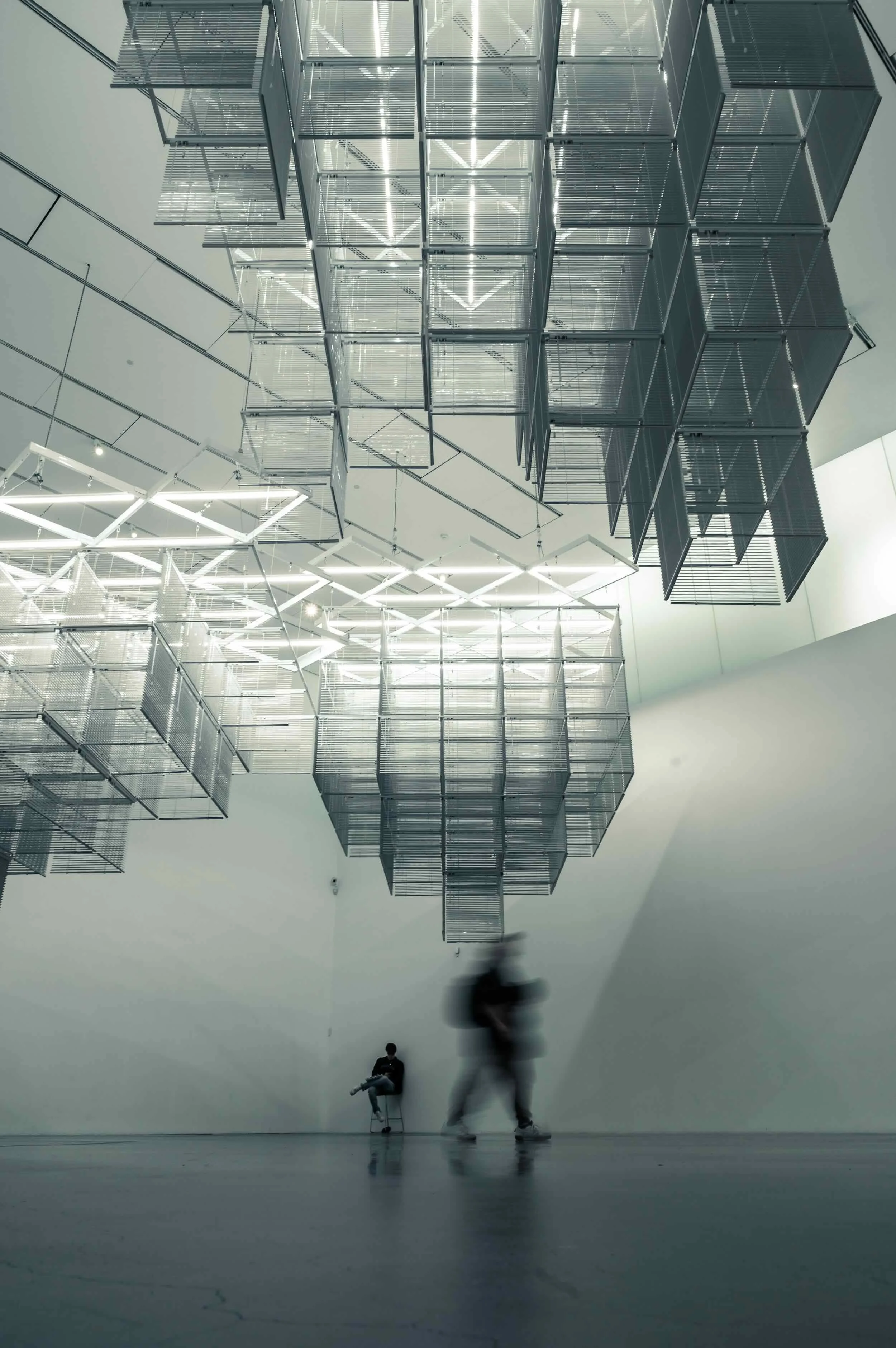 Modern art installation in a spacious gallery with suspended metallic cubes and geometric light fixtures, with blurry figures of people walking and sitting.