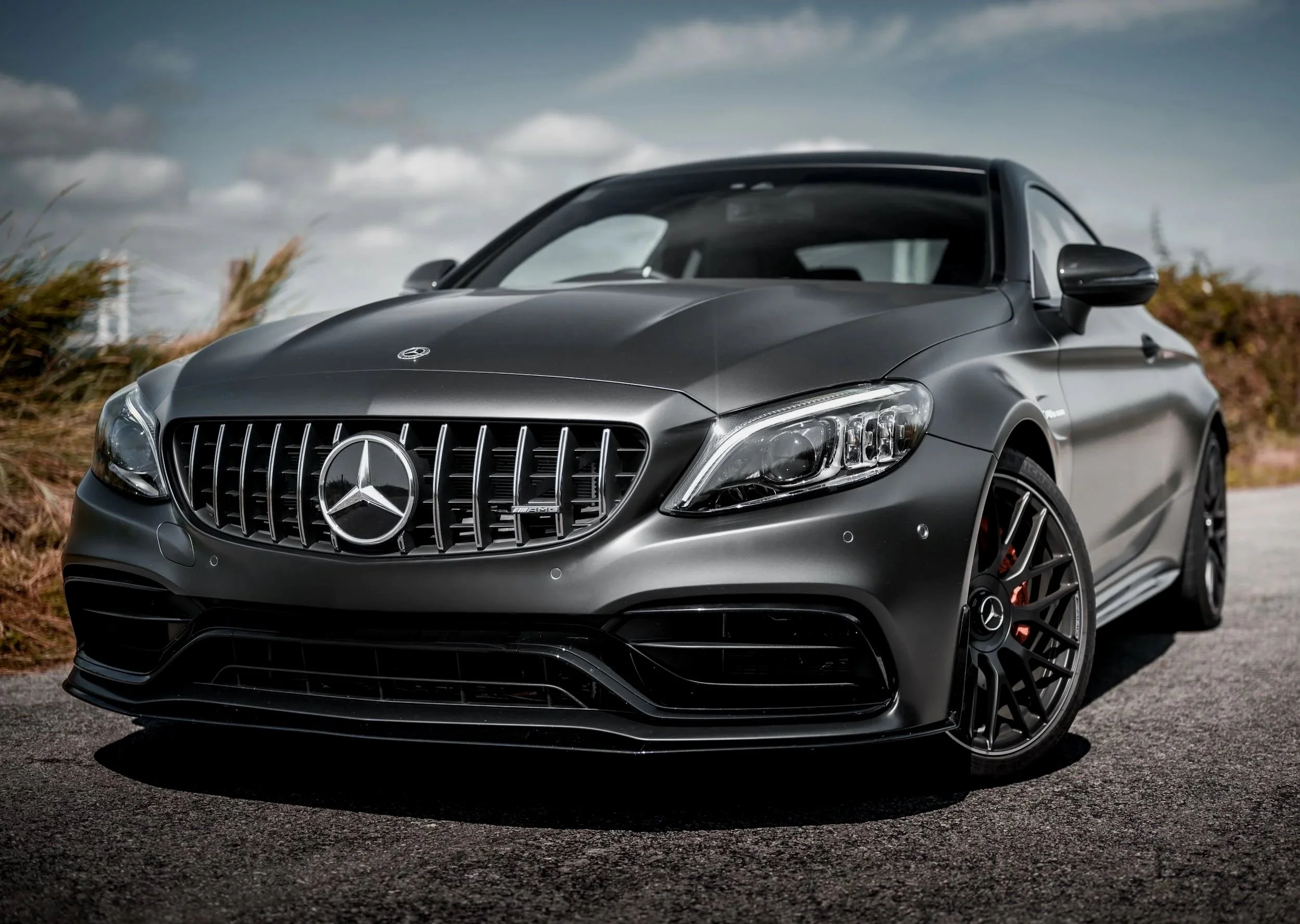 A sleek, dark gray Mercedes-Benz car with a prominent front grille and illuminated headlights parked outdoors on a paved surface with clouds in the sky.