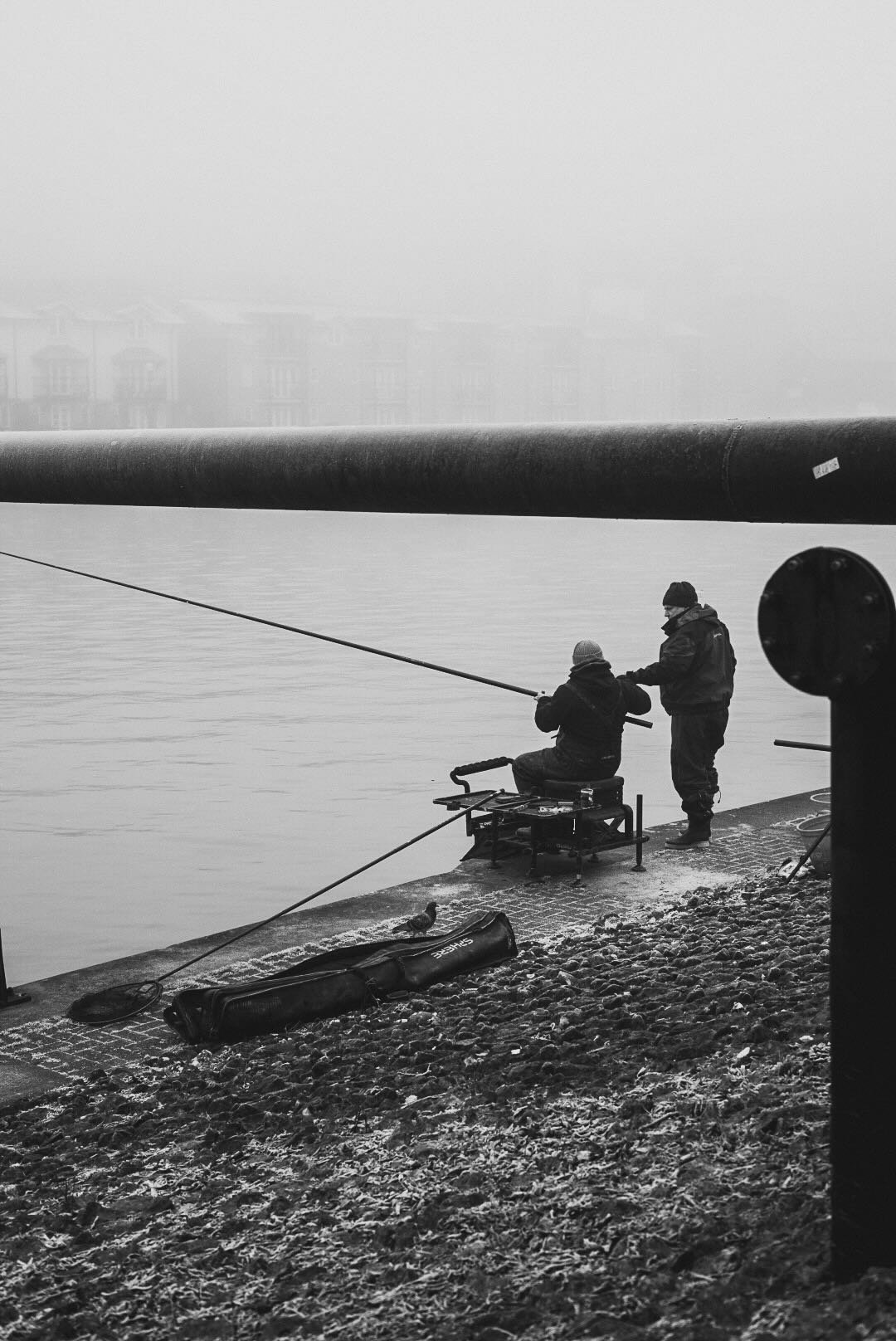 Two people fishing by the water's edge on a cold, foggy day. One person is sitting while the other is standing. A fishing rod and a tackle bag are on the ground.