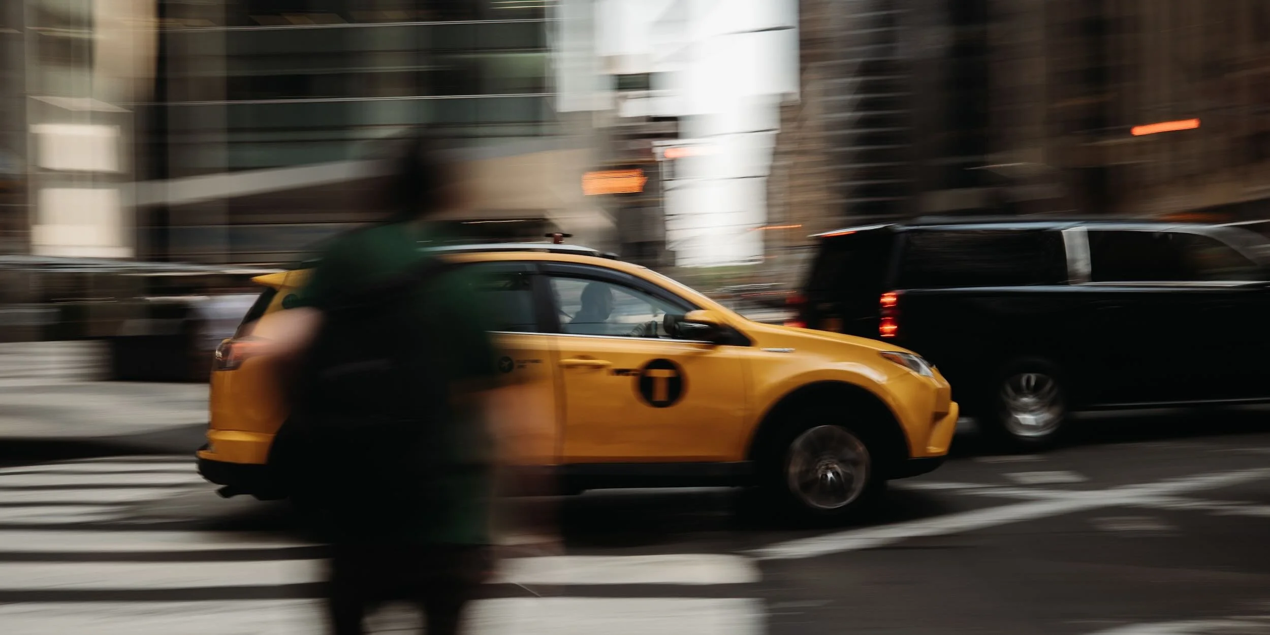 Blurry motion shot of a yellow taxi cab and other vehicles moving through a city street, with office buildings in the background.