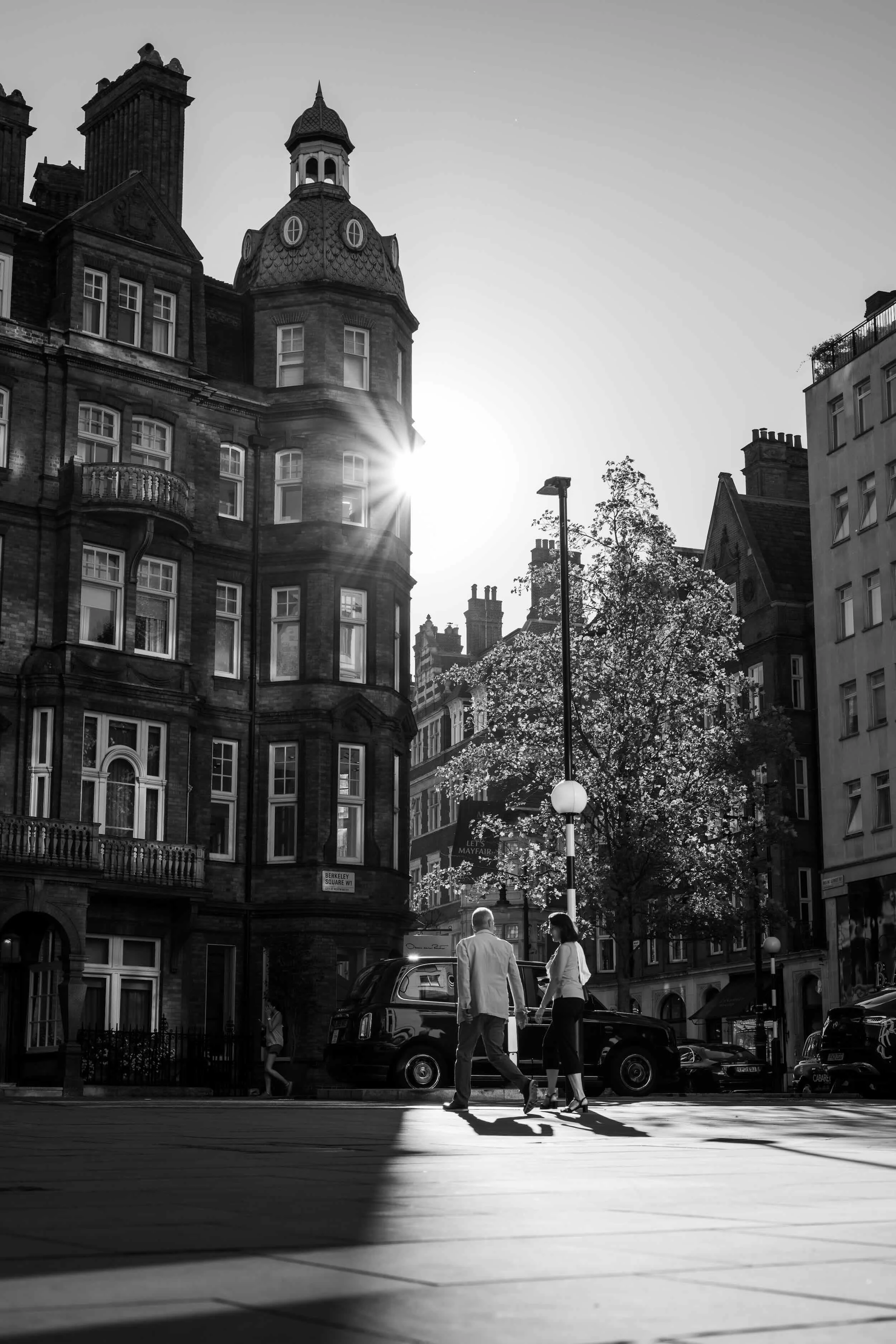 Black and white photo of people walking on a city street with historic buildings and cars parked along the curb, the sun shining through the buildings.