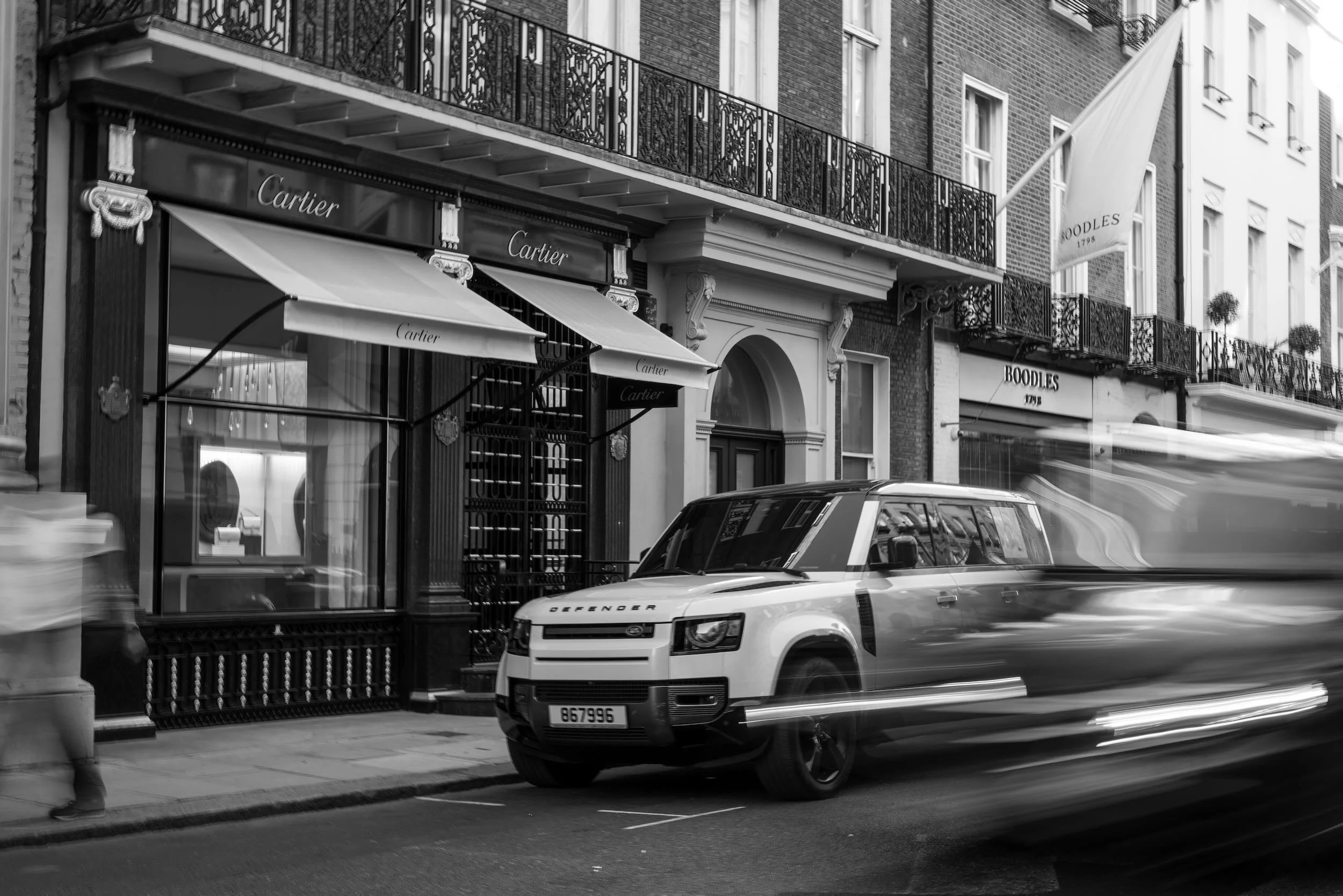 A black and white photograph of a city street with luxury and boutique storefronts. The storefronts include Cartier with awnings and ornate window details, and Boodles with a sign indicating 1798. A Land Rover Defender is parked on the street, and th