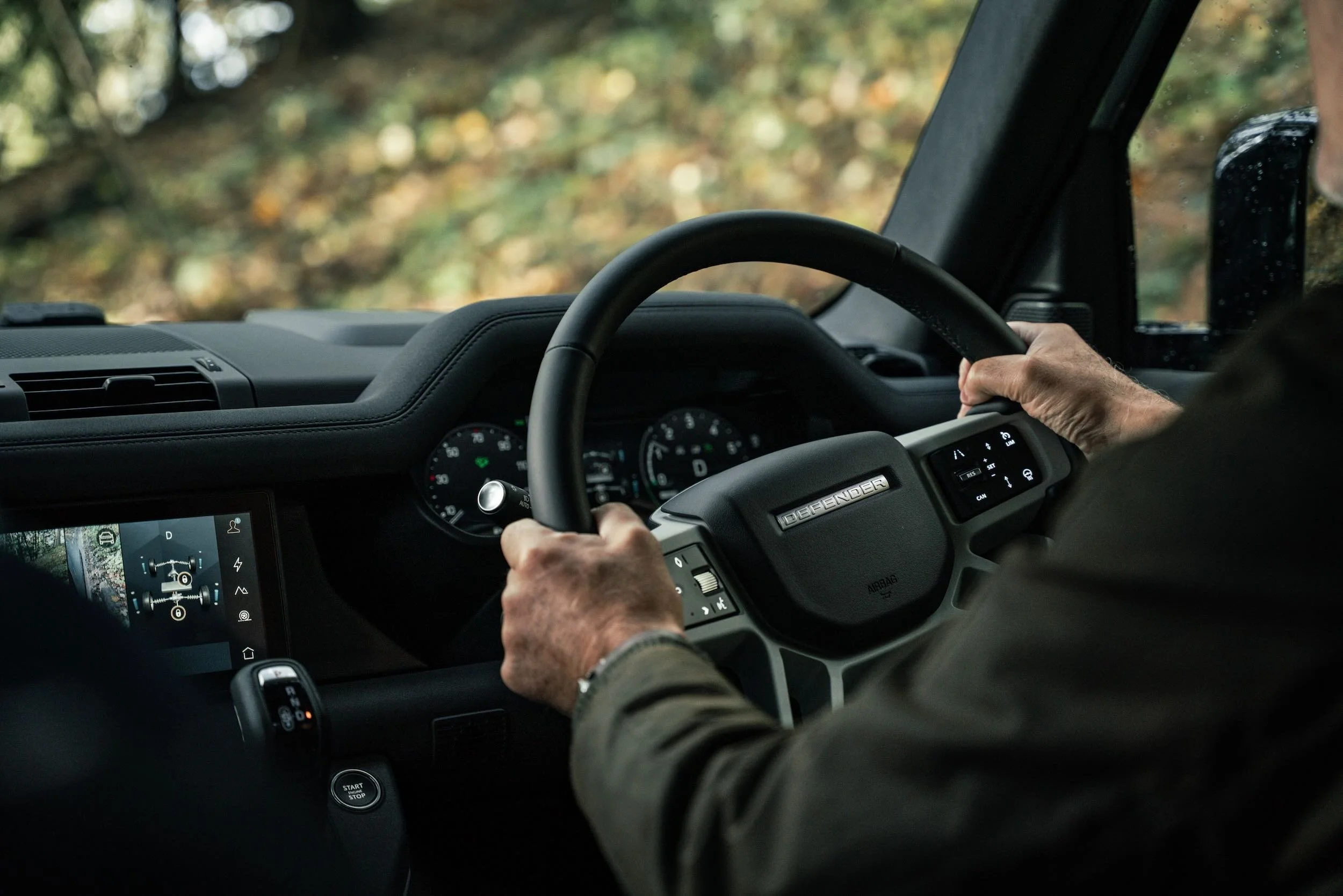 Interior of a vehicle with a person driving, showing hands on the steering wheel and dashboard, with a blurred outdoor background visible through the window.