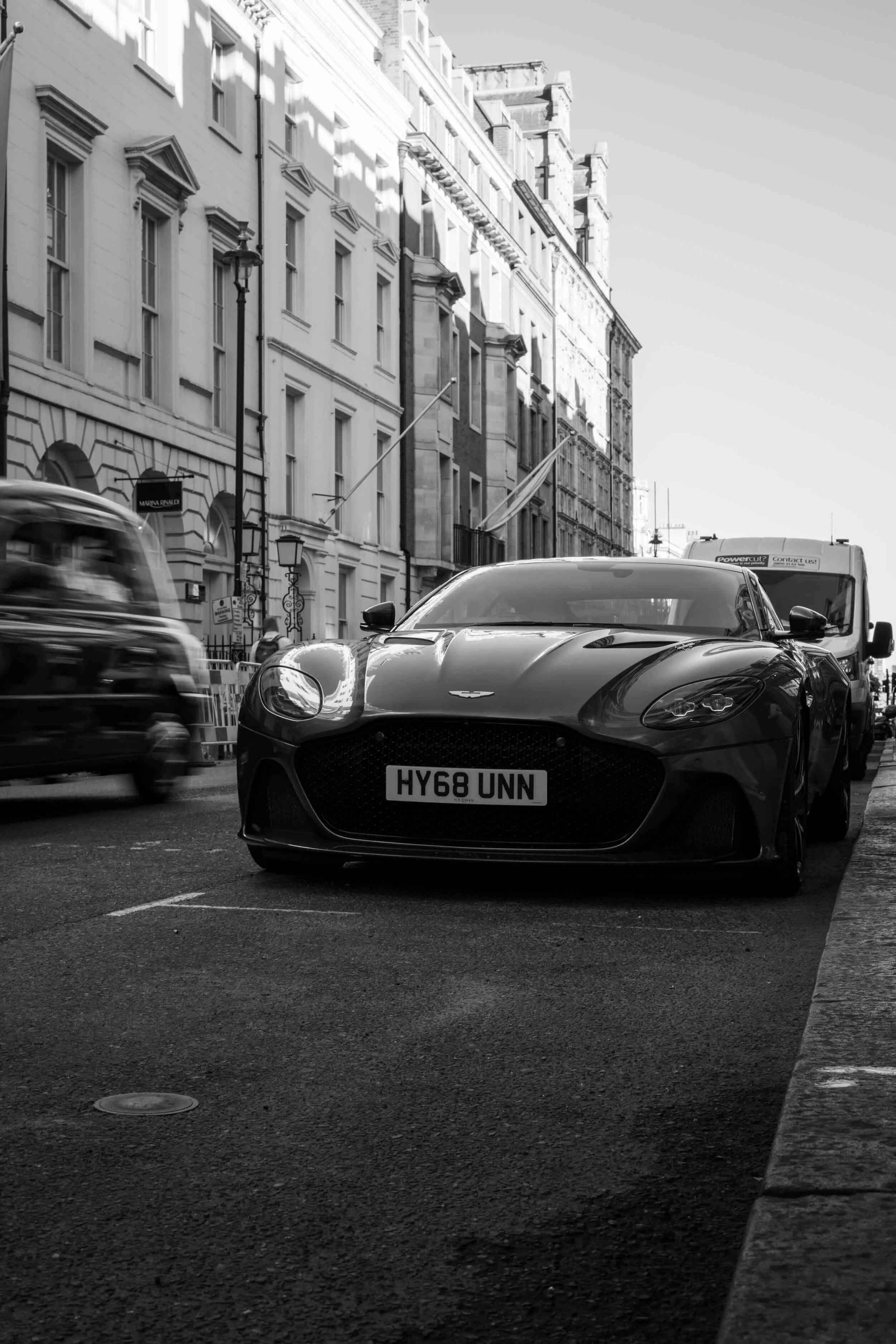 A black sports car with a UK license plate 'HY68 UNN' parked on a city street, with blurred vehicles passing by and historic buildings with ornate facades in the background.