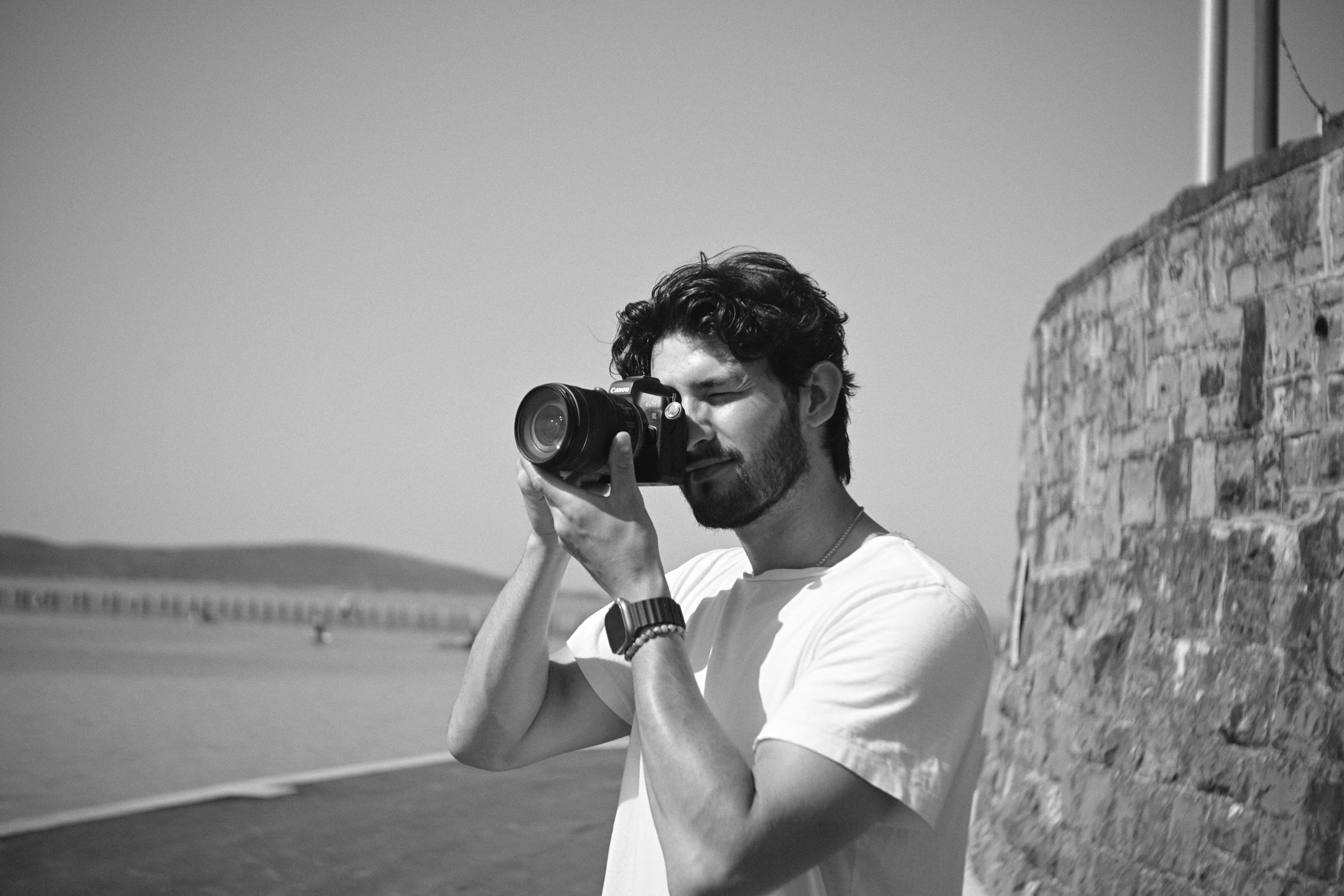 A man with dark curly hair and a beard looking through the viewfinder of a camera, standing outdoors near a stone wall on a sunny day.