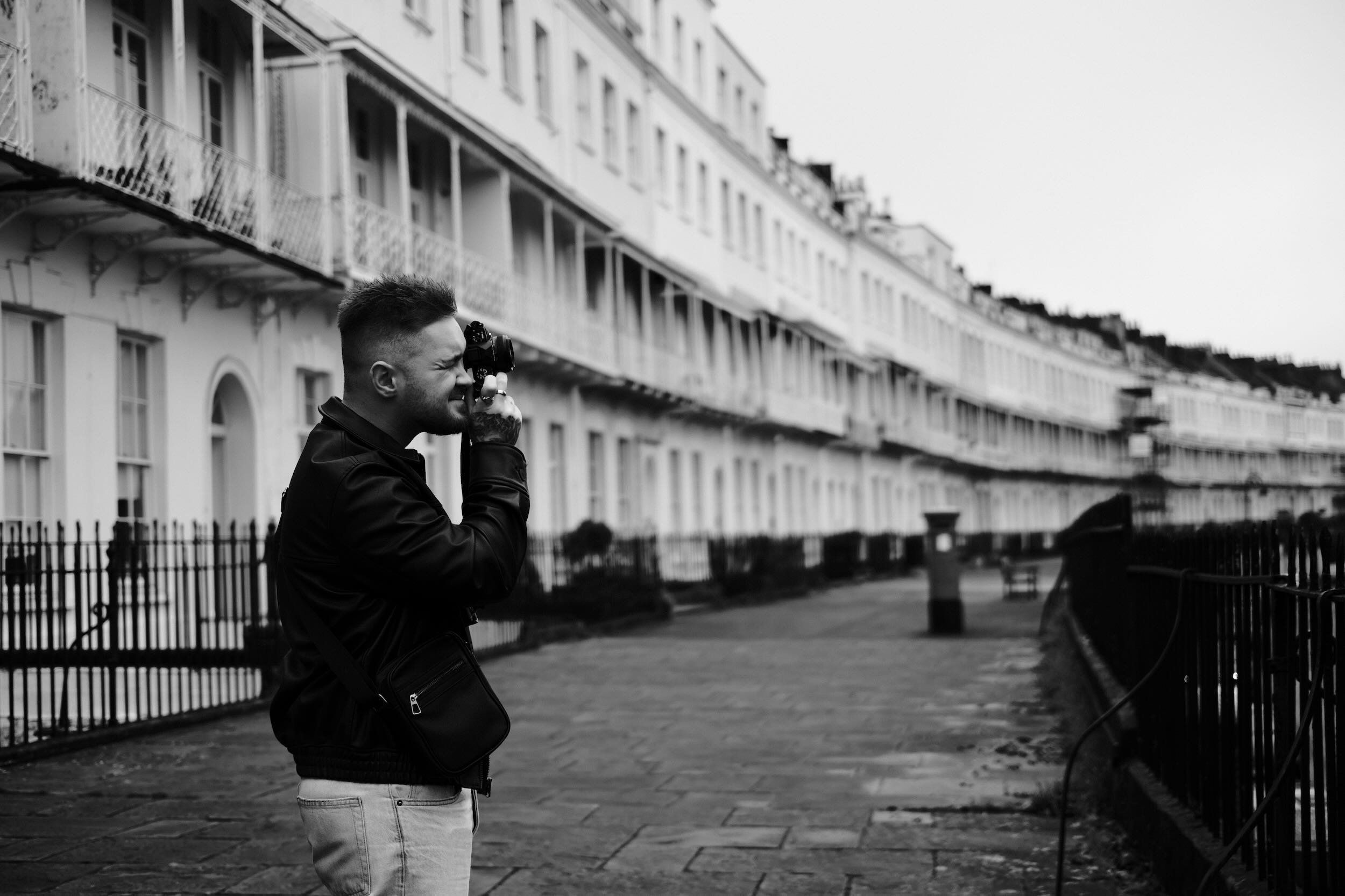 A man in a black jacket and light jeans is taking a photo with a camera on a city street. In the background, there are multi-story white residential buildings with balconies and black fences.