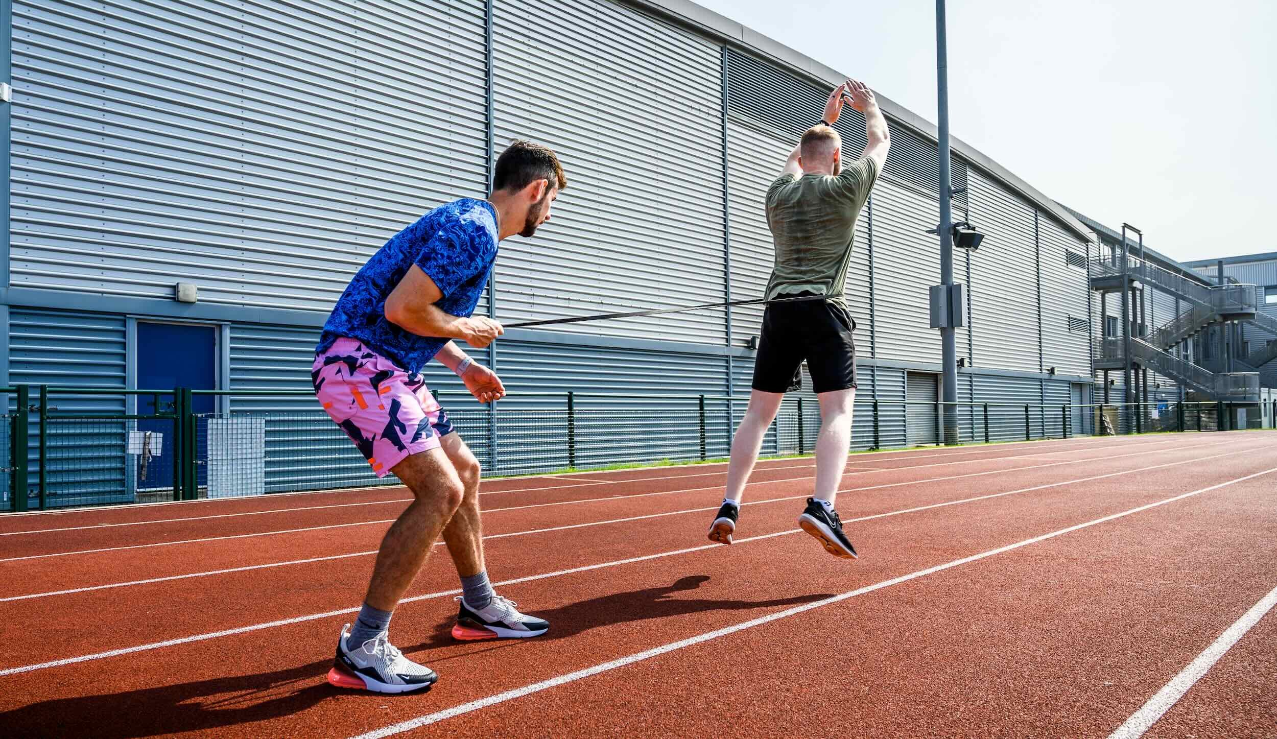 Two young men on a running track, one jumping and the other jumping rope. The background shows a large industrial-style building with metal siding.