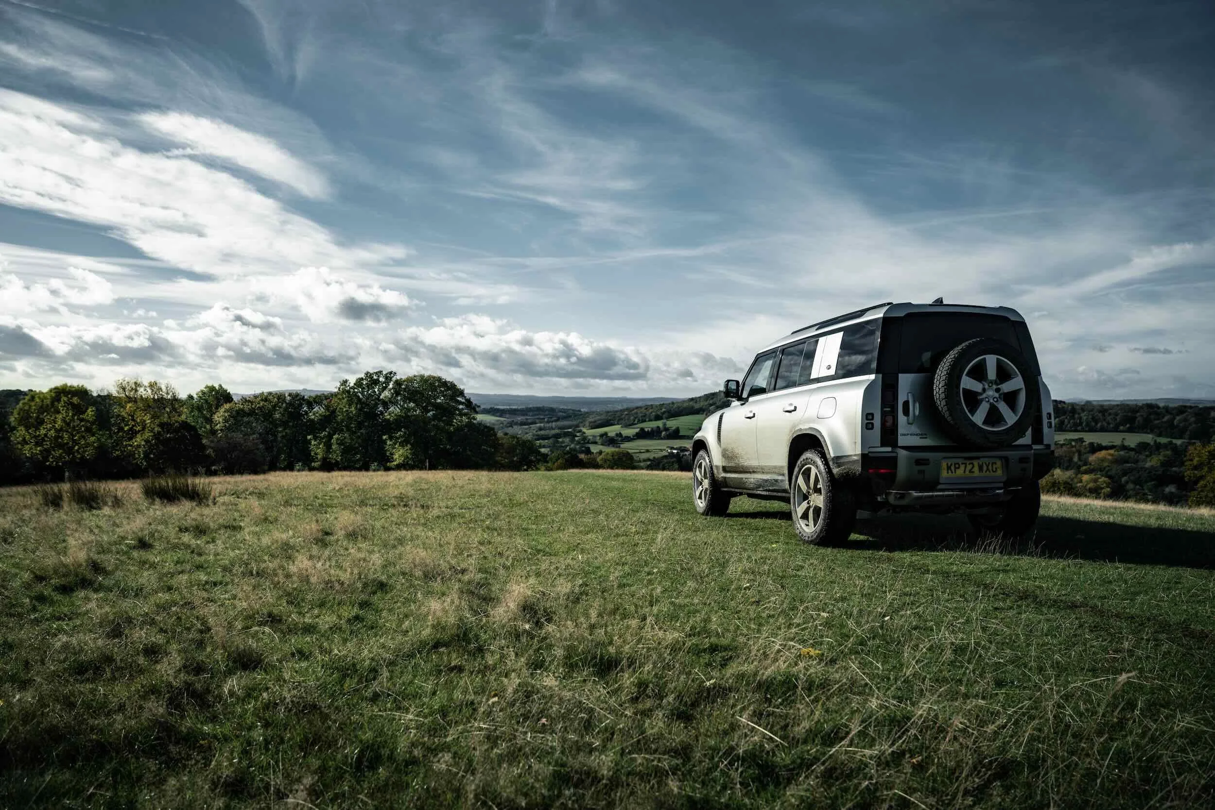 A silver SUV parked on a grassy field with trees and hills in the background under a cloudy sky.