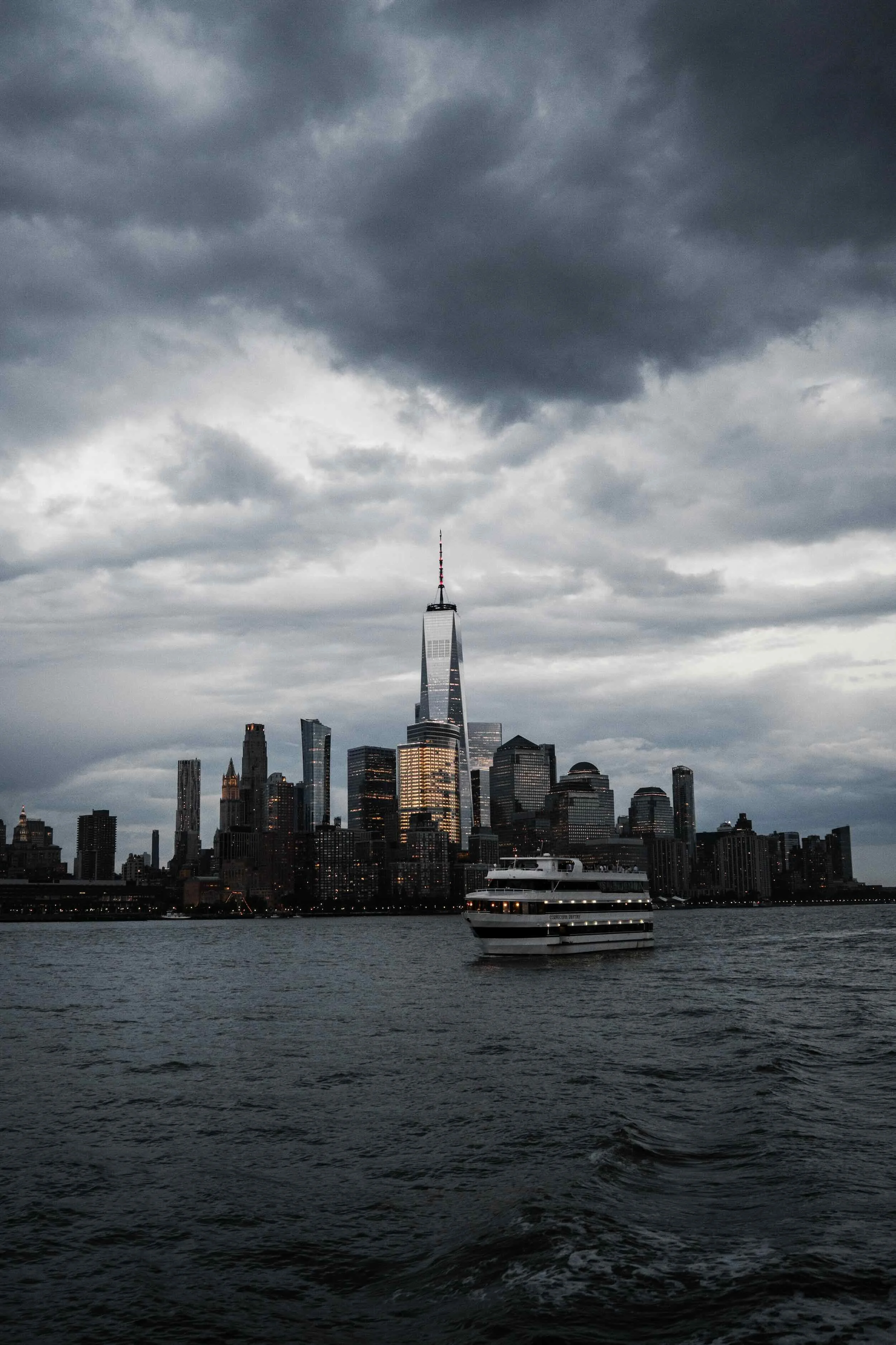 View of a city skyline with tall buildings and skyscrapers under a cloudy sky, with a boat sailing across the water in the foreground.