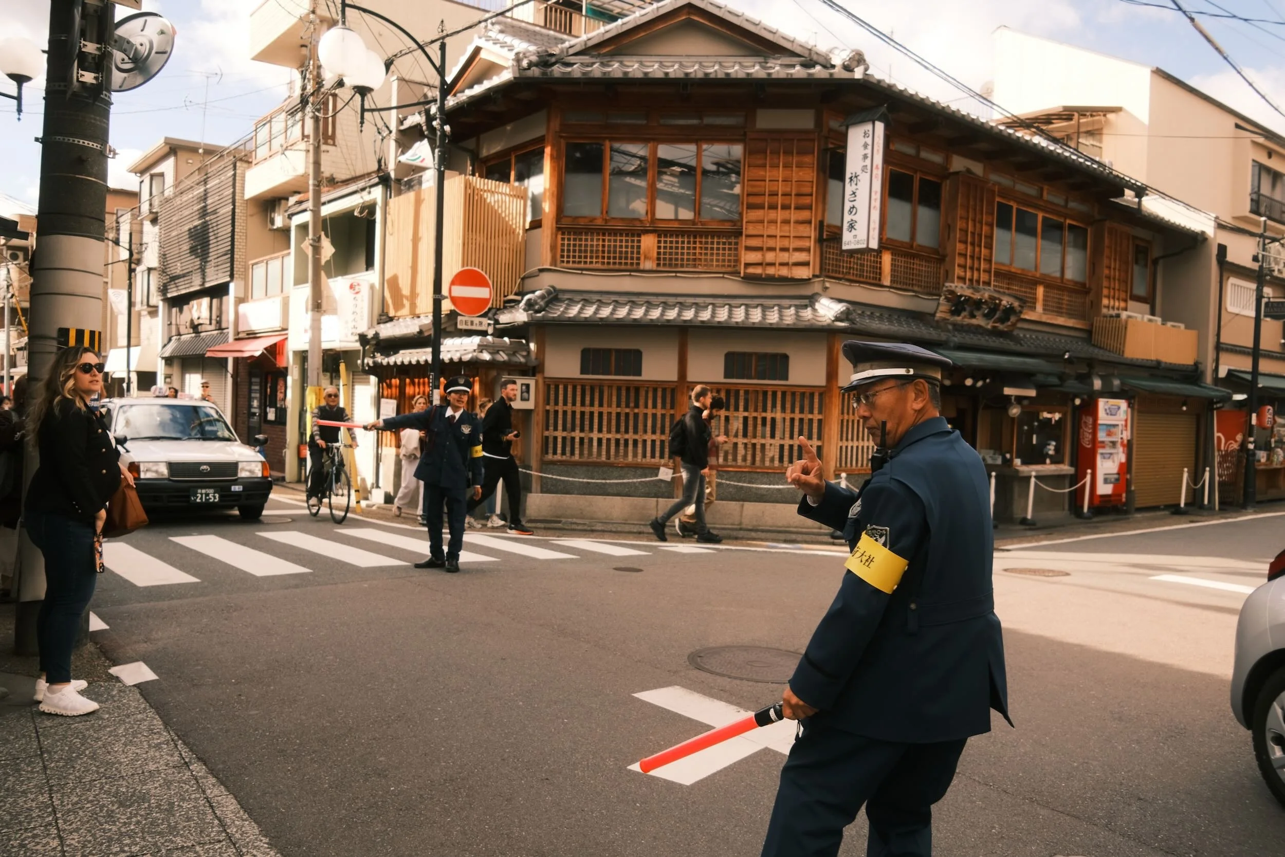 A crosswalk in a busy urban street in Japan with pedestrians and a police officer directing traffic. Traditional wooden buildings with signs and shops line the background, and a woman in sunglasses waits at the crosswalk.