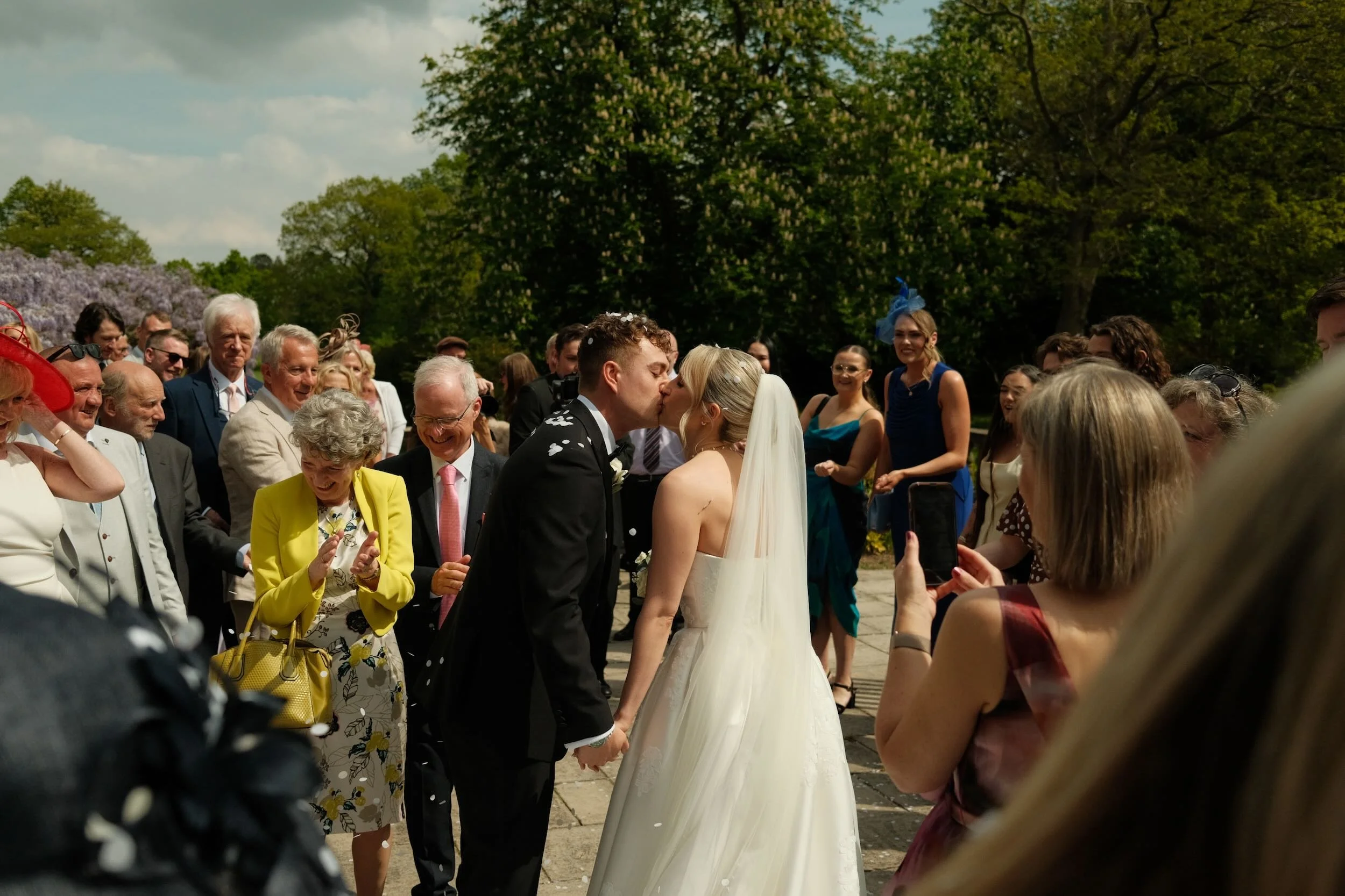 A bride and groom kiss during their outdoor wedding ceremony as guests watch and take photos around them.