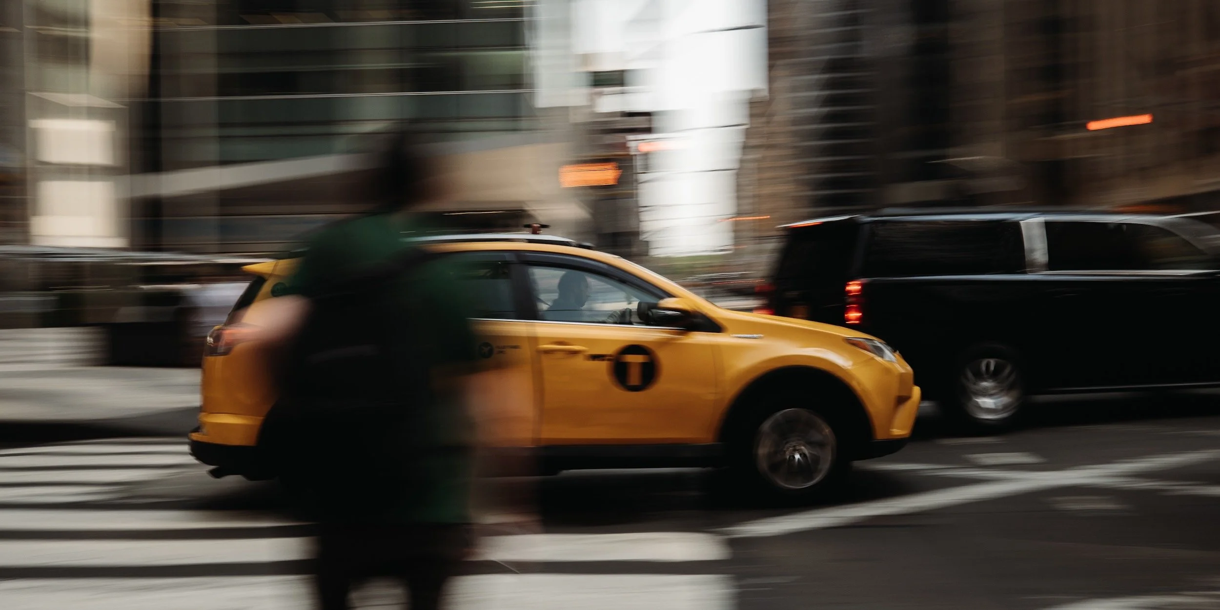 A blurred city street scene with a yellow taxi and a black van moving past pedestrians, with buildings in the background.