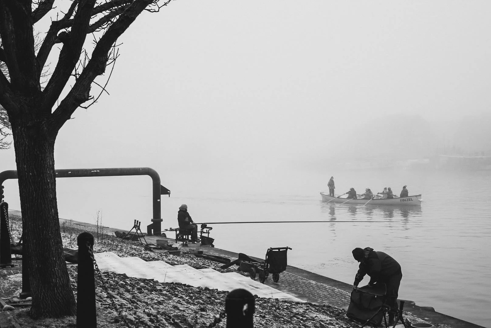 People by the water's edge on a cold day, with one person fishing and another bending down near a bicycle or equipment, while a boat carrying several passengers is on the water, enveloped in fog.