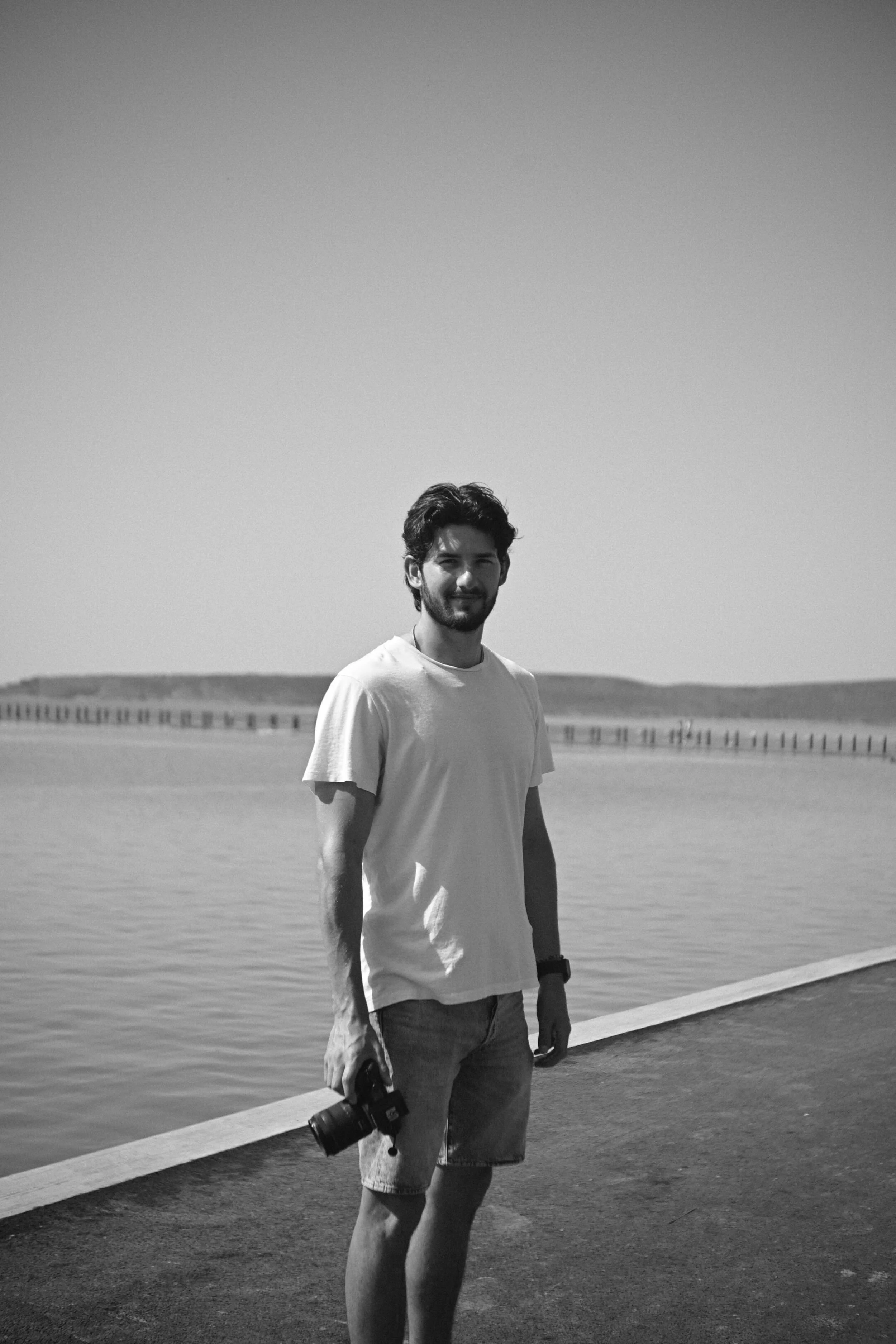 A young man with dark hair and beard, wearing a white T-shirt and shorts, standing by a body of water on a cloudy day, holding a camera, with a pier in the background.
