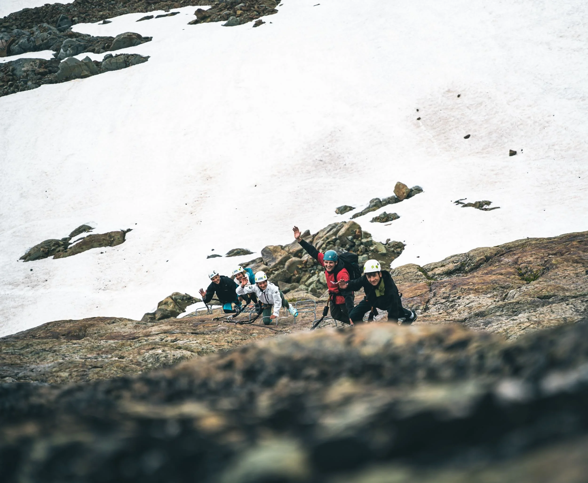 A group of people climbing a rocky mountain trail with snow patches, wearing helmets and outdoor gear, with greenery and snow in the background.