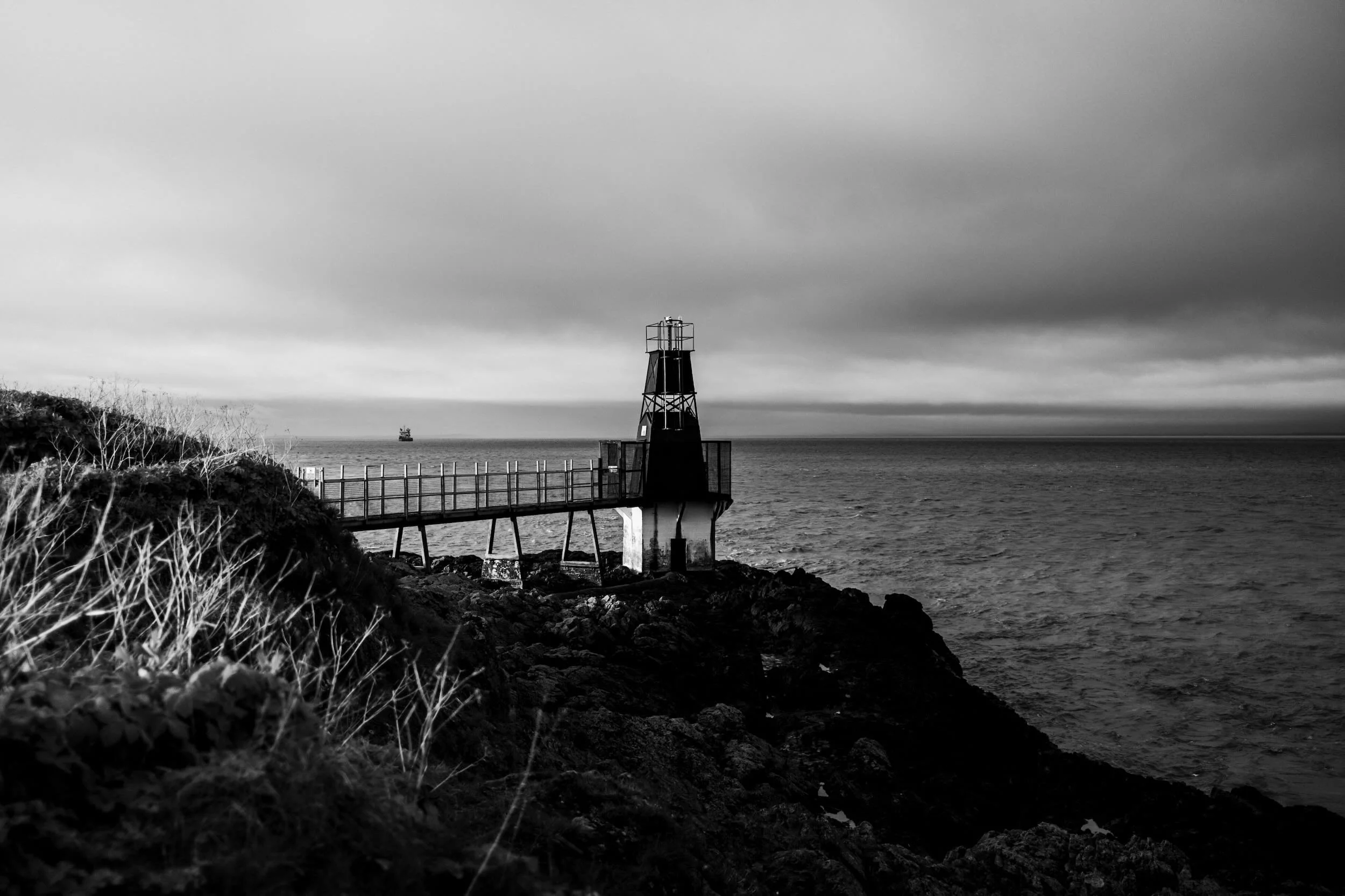 A black and white photo of a lighthouse on a rocky coast with a railing, with the ocean and a ship in the background under a cloudy sky.