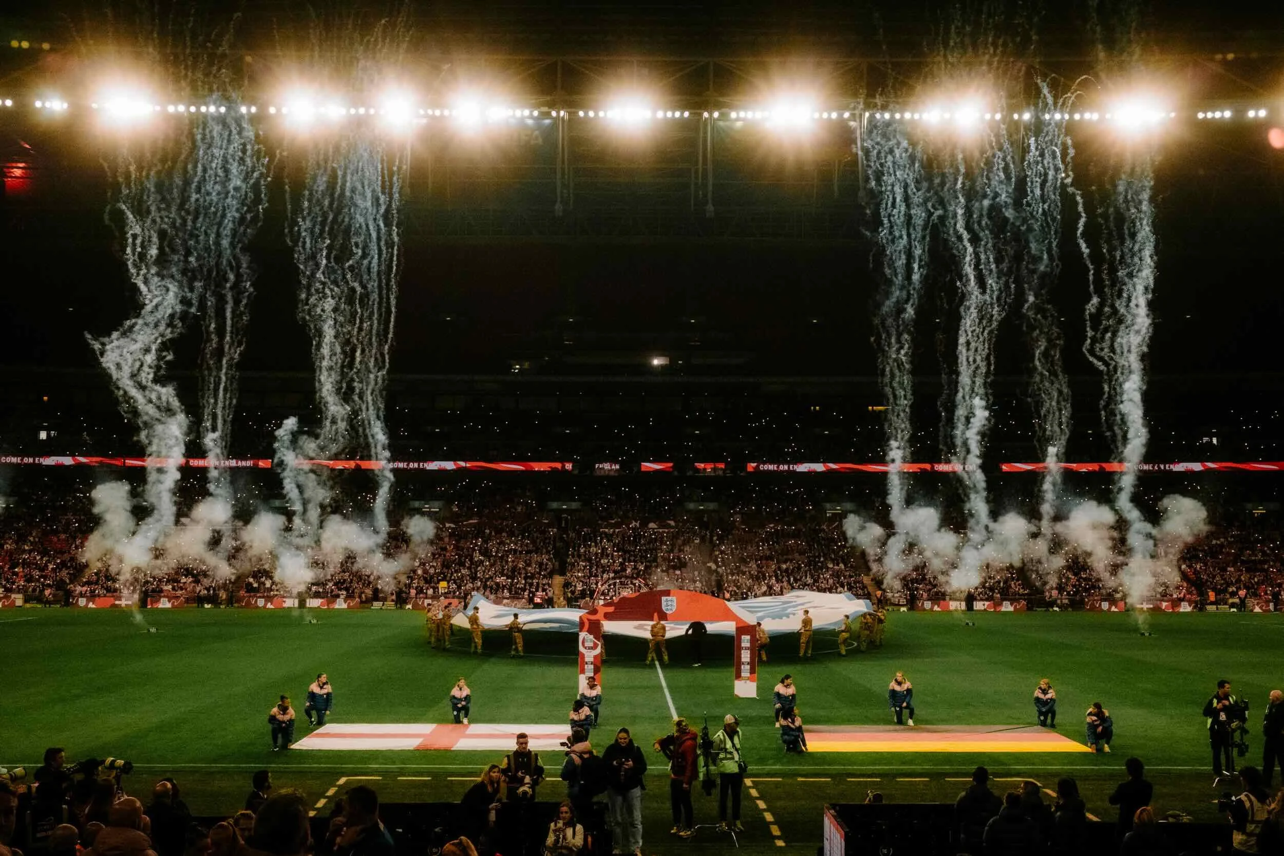 A stadium filled with spectators, with bright stadium lights and smoke on the field. People are gathered around, and some are holding a large flag of England. Fireworks or pyrotechnics are being launched in the background.