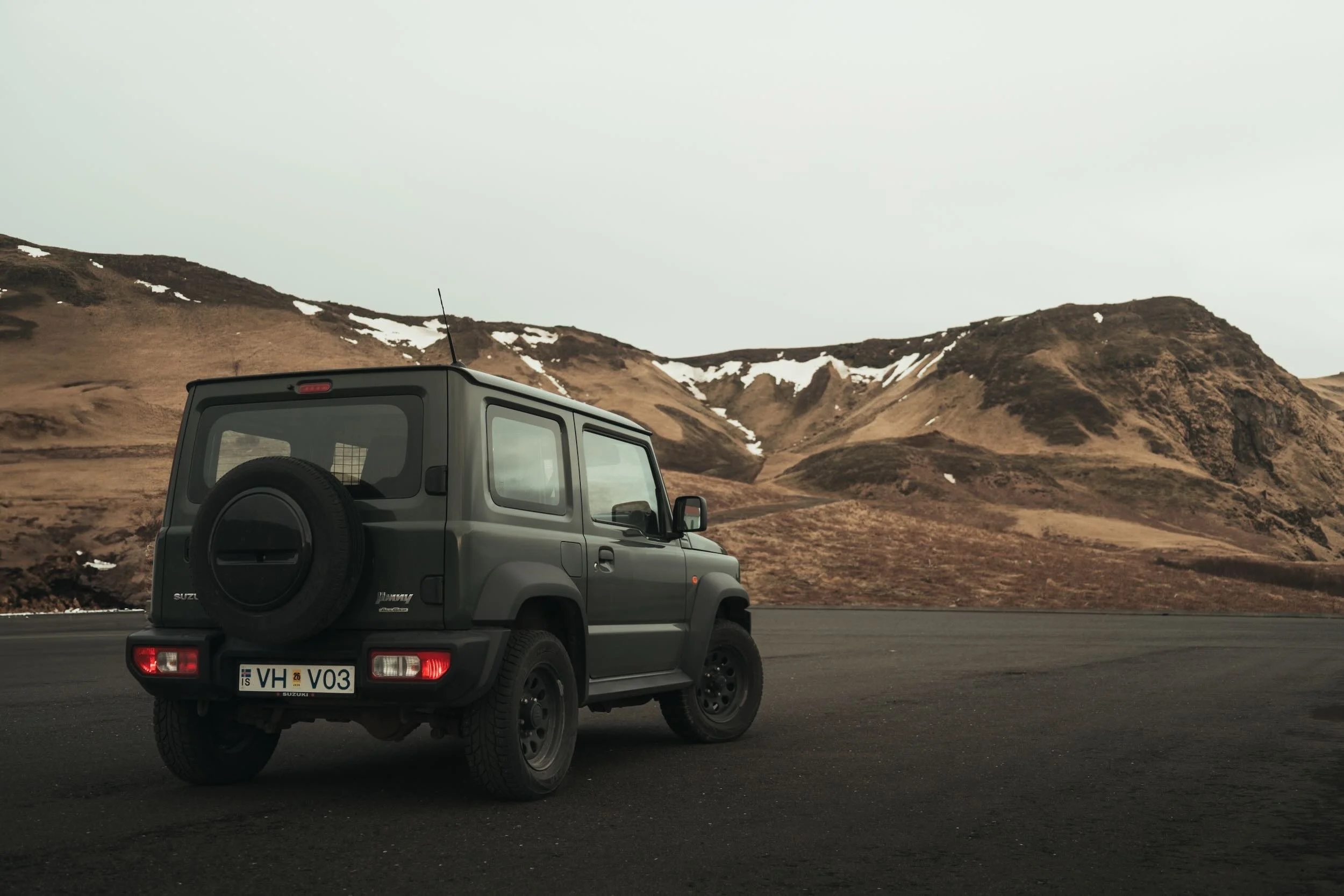 A gray Suzuki Jimny SUV parked on the side of a road with rocky, snow-capped mountains in the background.