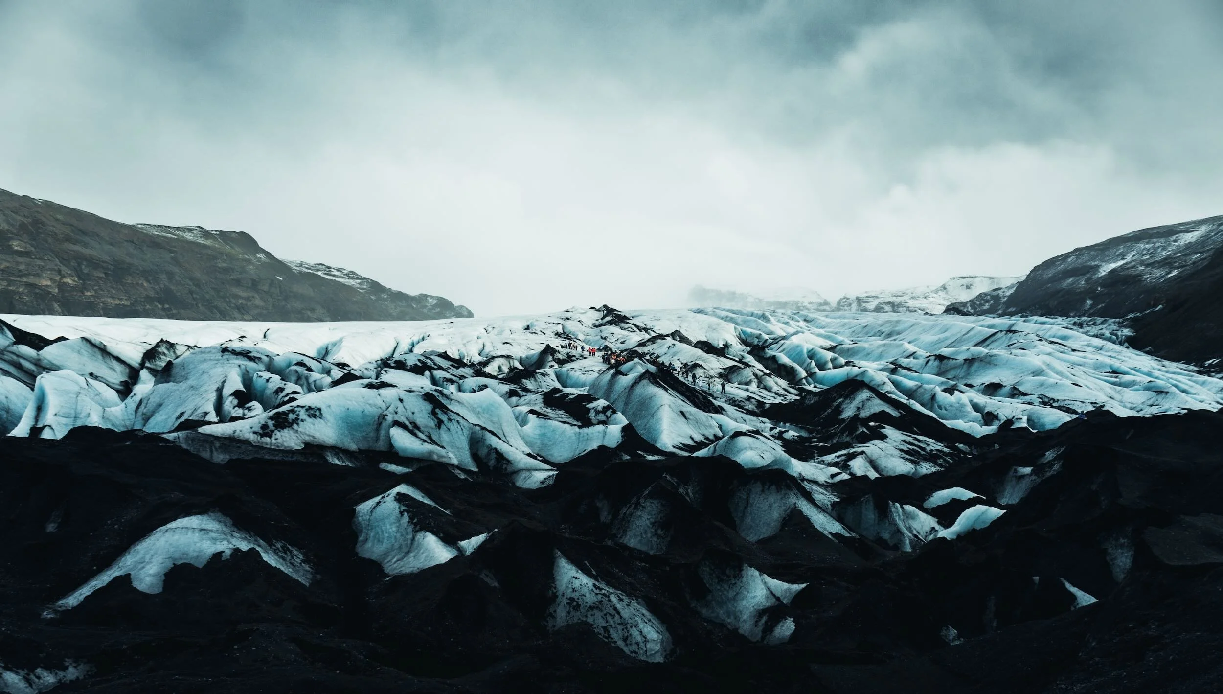 View of a glacier with blue ice and black volcanic ash streaks, surrounded by mountain terrain under a cloudy sky.