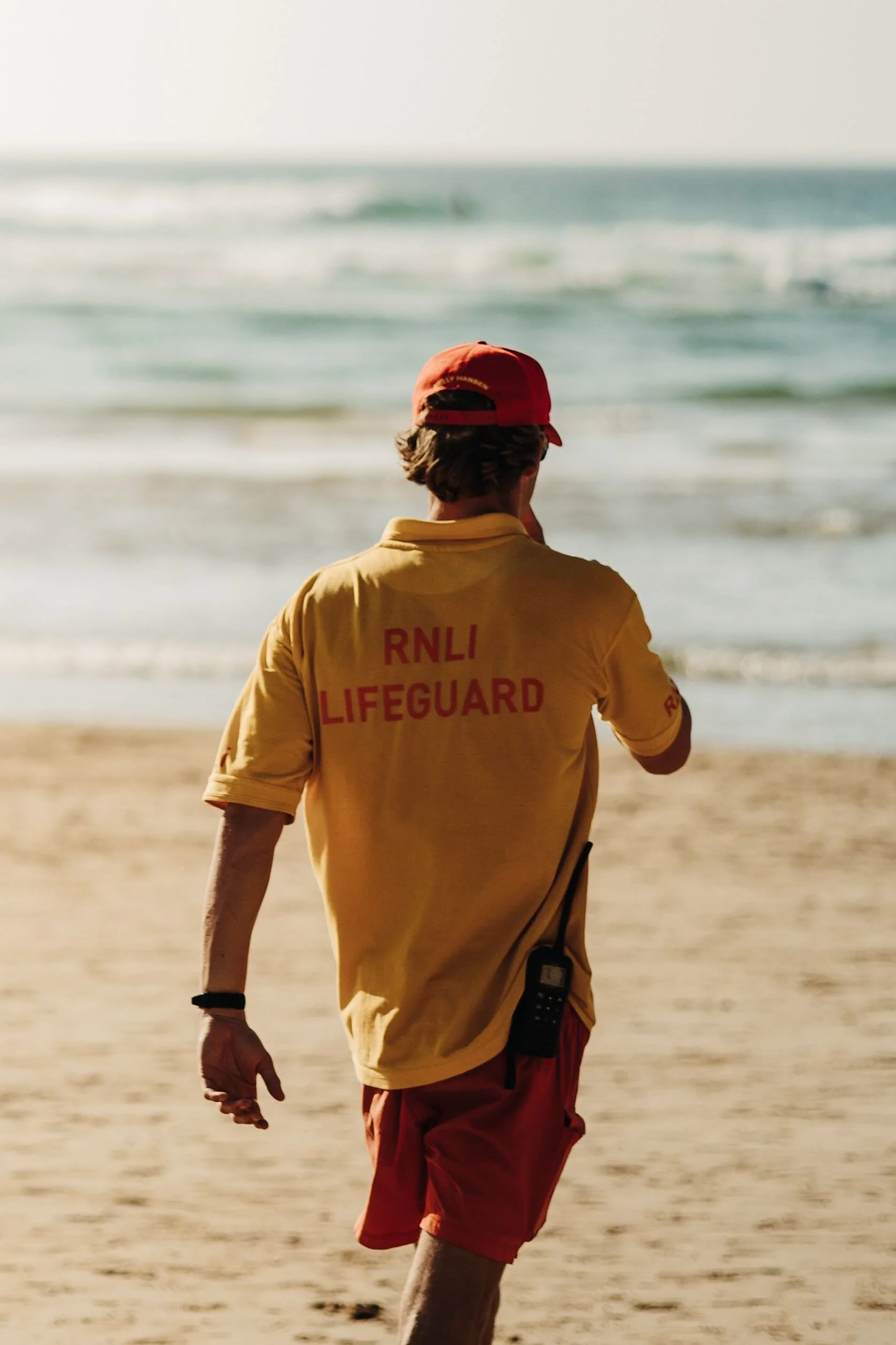 RNLI lifeguard wearing a yellow shirt and red cap walking along the beach facing the ocean.