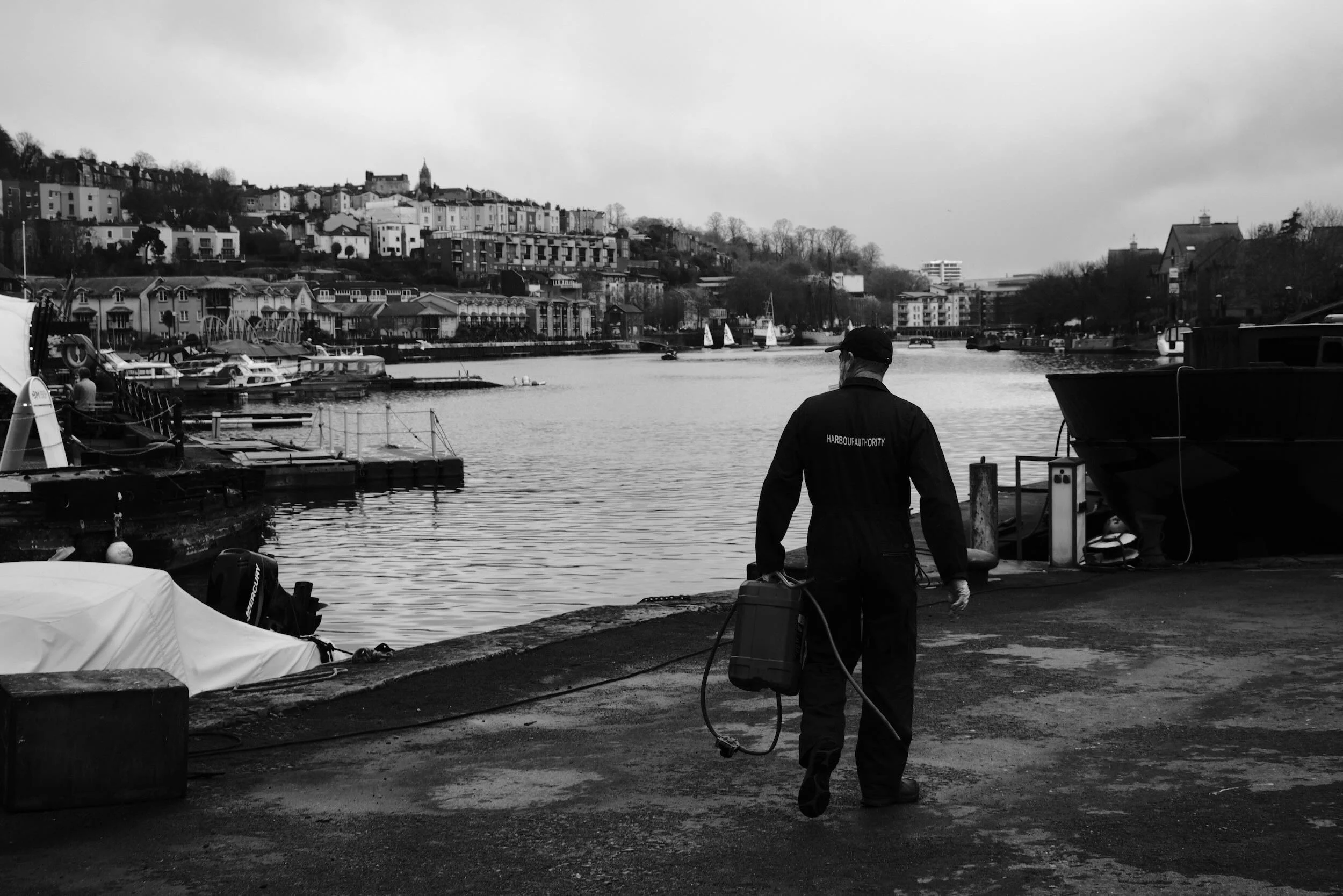 A man walking by a harbor with boats, buildings on a hillside, and sailboats on the water in the background, in black and white.