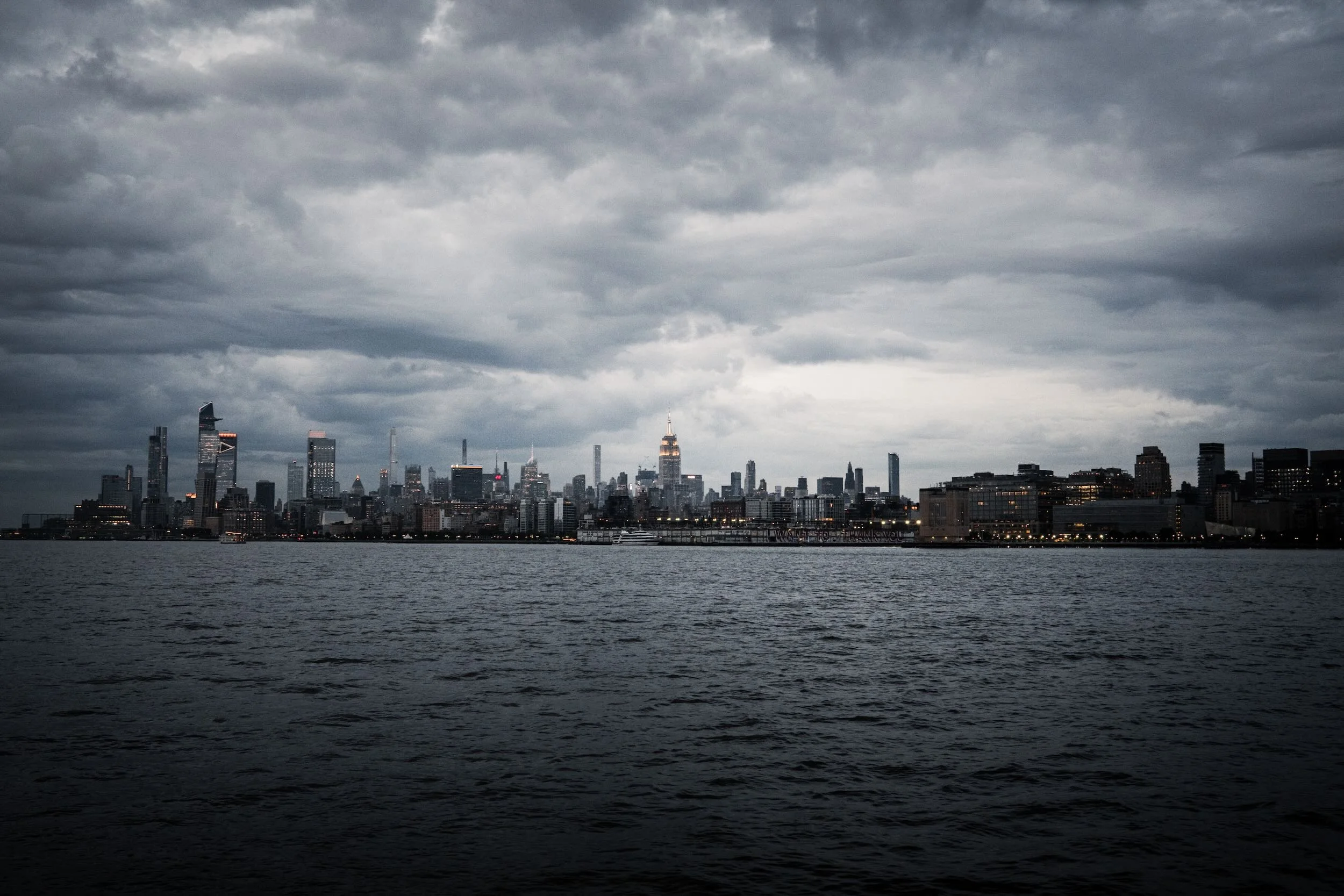 A city skyline with tall buildings under a dark, cloudy sky, viewed from across a body of water.