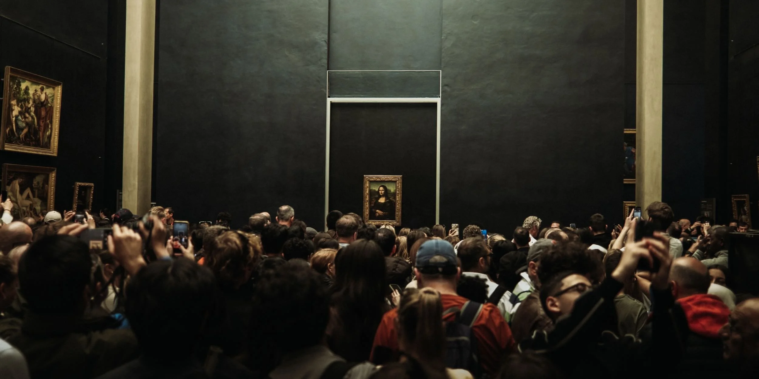 A large crowd of people gathered in an art museum, facing a massive black wall with the Mona Lisa painting displayed in the center.