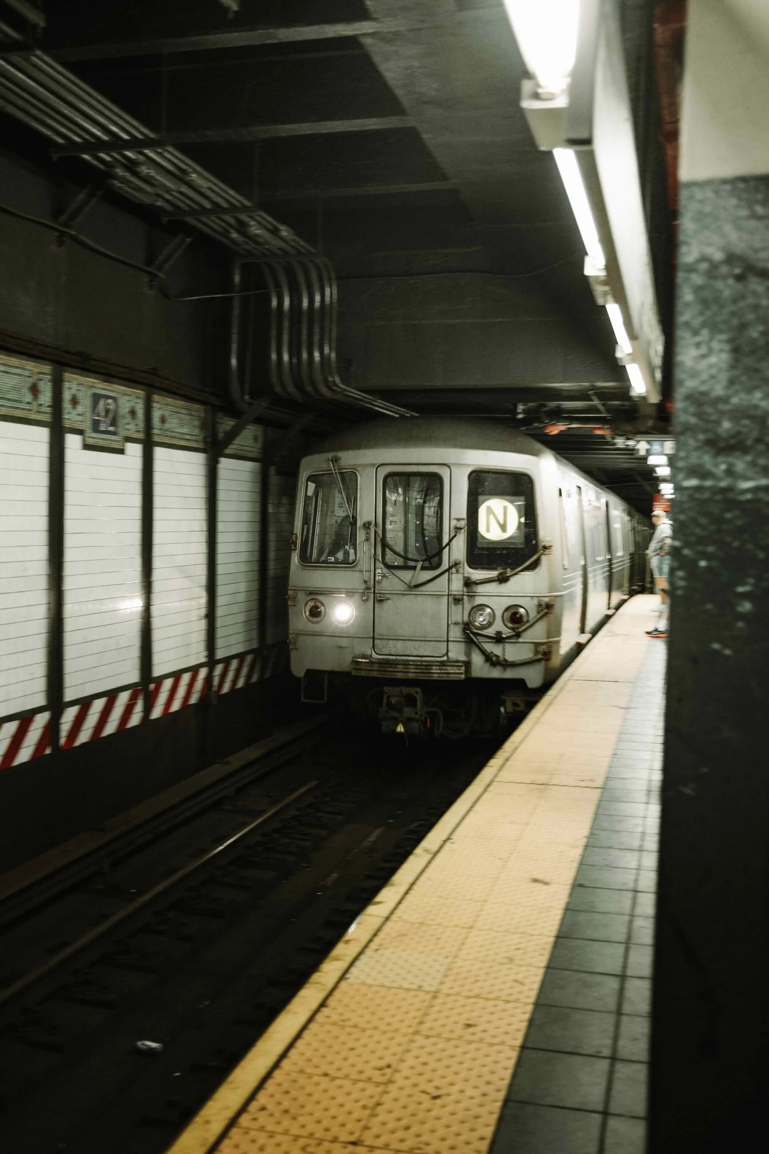 A subway train with an 'N' logo on the front is stopped at the platform in an underground station. The platform has a yellow tactile paving strip along the edge, and there are some passengers waiting further down the platform. The ceiling is dark wit