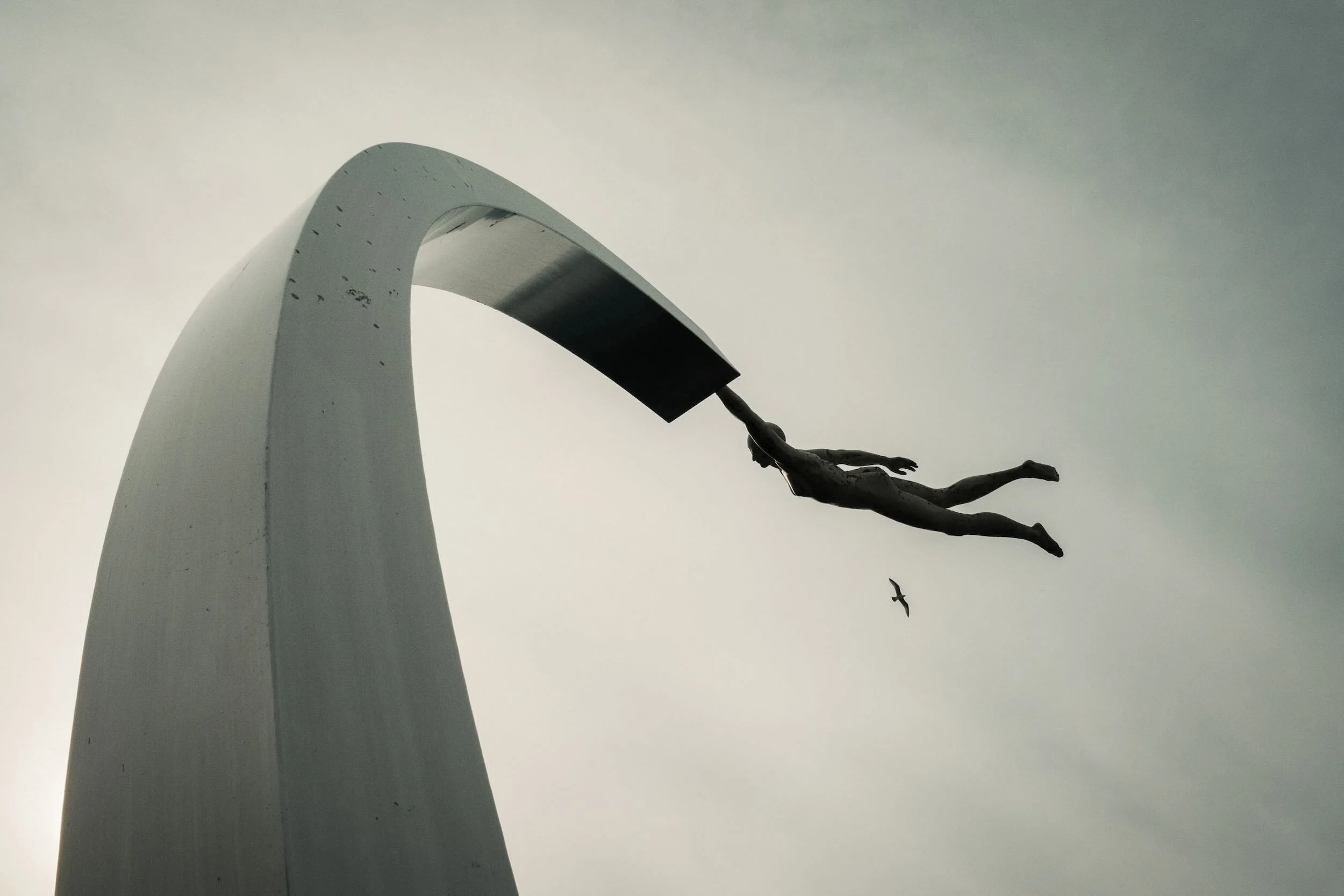 A sculpture of a human figure floating horizontally, holding onto a rope attached to a curved, white architectural structure, with birds flying in the sky in the background.