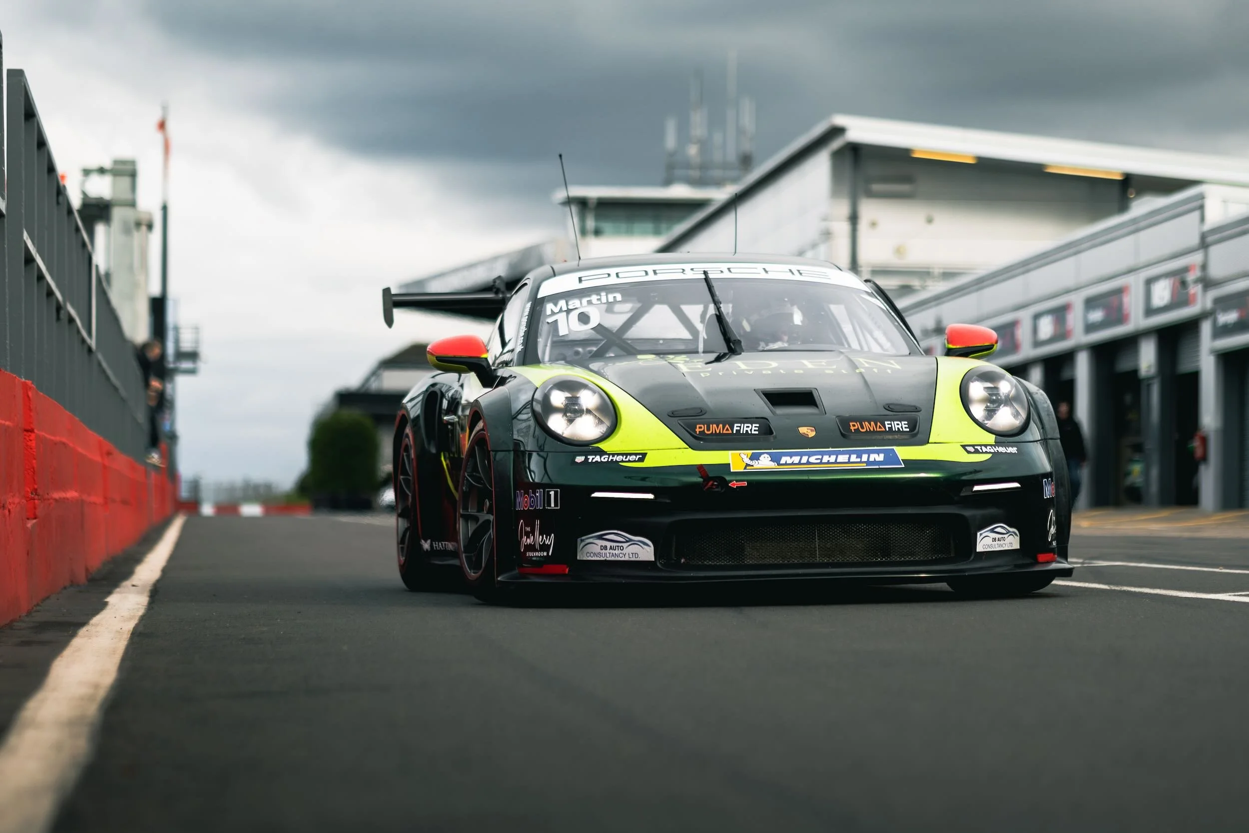 A racing car on a track with a dark, cloudy sky in the background. The car has a black and green color scheme with sponsor logos, prominent headlights, and a rear wing.