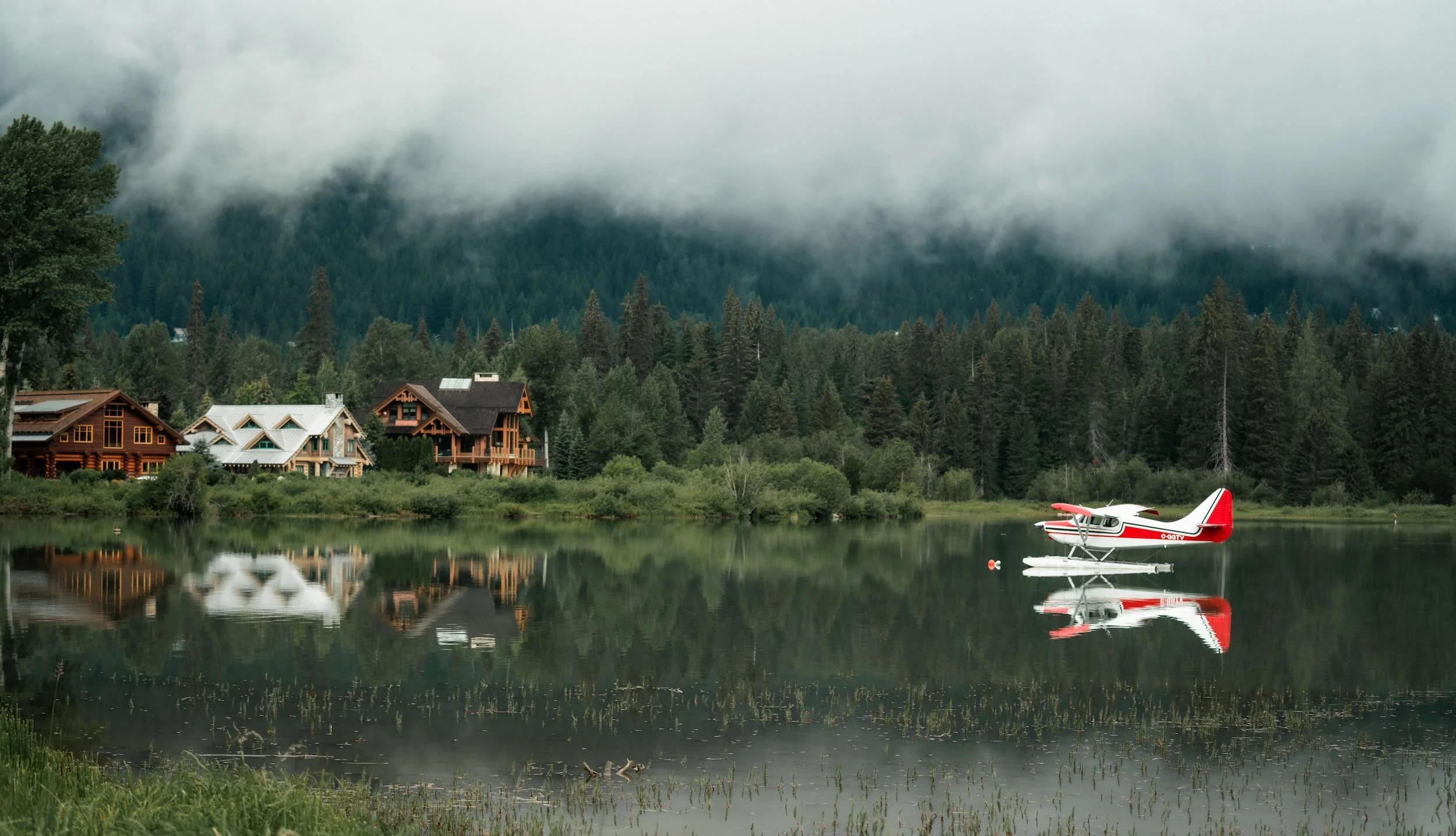 Calm lake with a small red and white seaplane floating on the water, surrounded by green trees and wooden houses, with mountains and fog in the background.