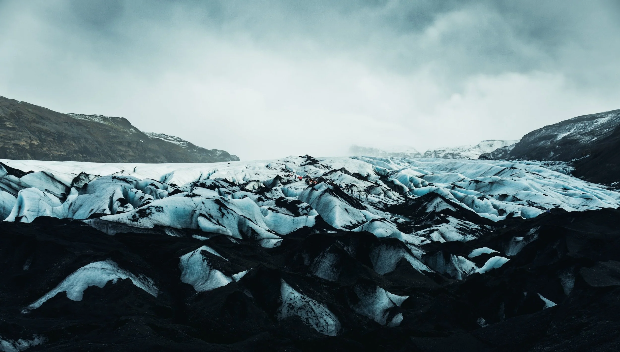 A large glacier with blue and white ice, with dark volcanic ash on parts of the ice, nestled between mountains under a cloudy sky. There are small groups of people visible on the glacier.