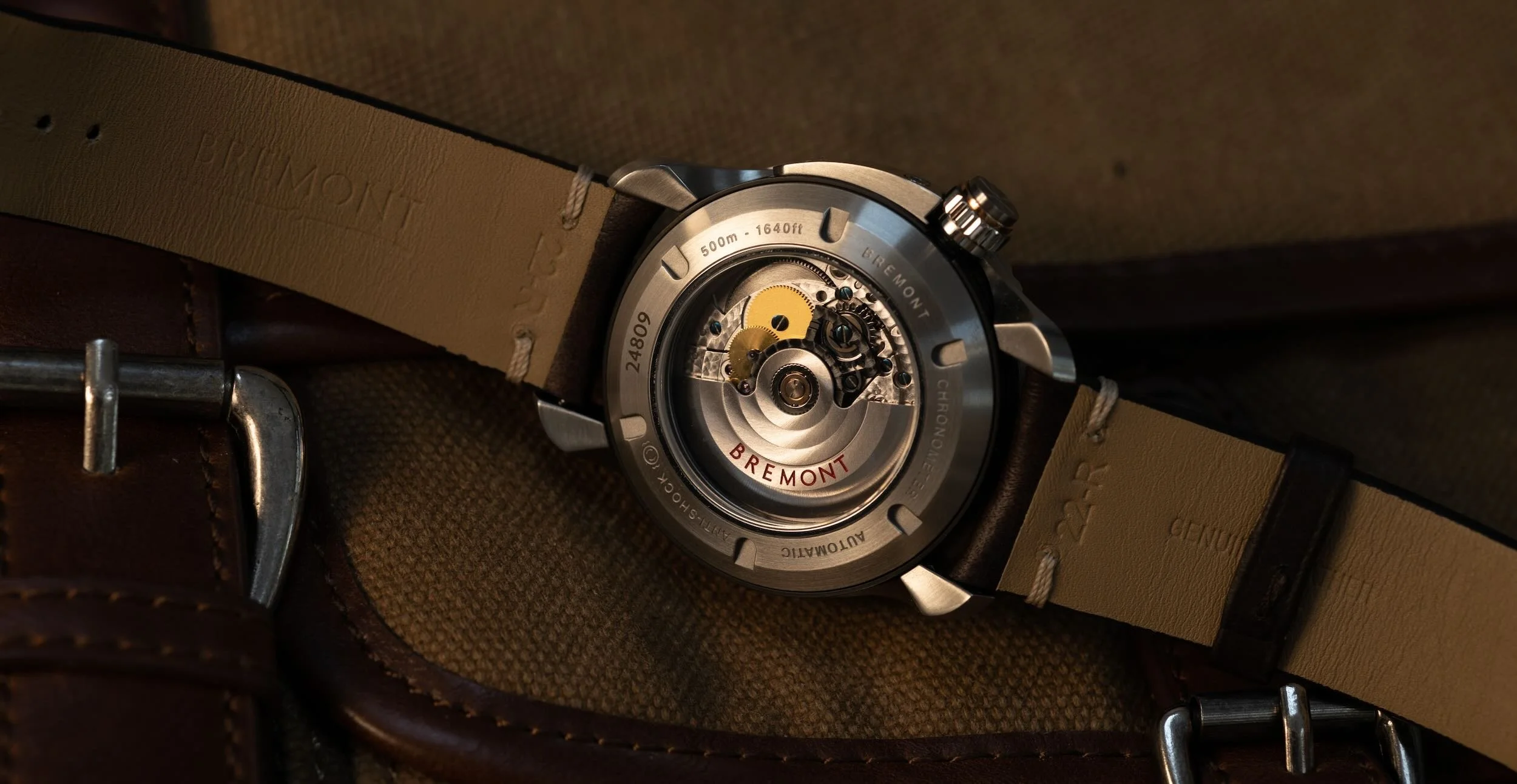 Close-up of a Bremont wristwatch with a transparent case back, revealing the mechanical movement inside, resting on a textured surface with a brown leather strap.