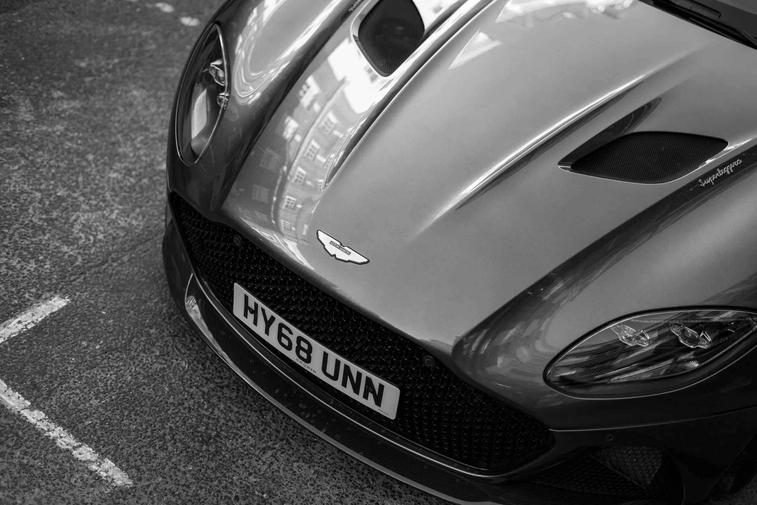 Front view of a sleek, silver Aston Martin sports car parked on a textured asphalt surface, showcasing the car's grille, headlight, and badge.