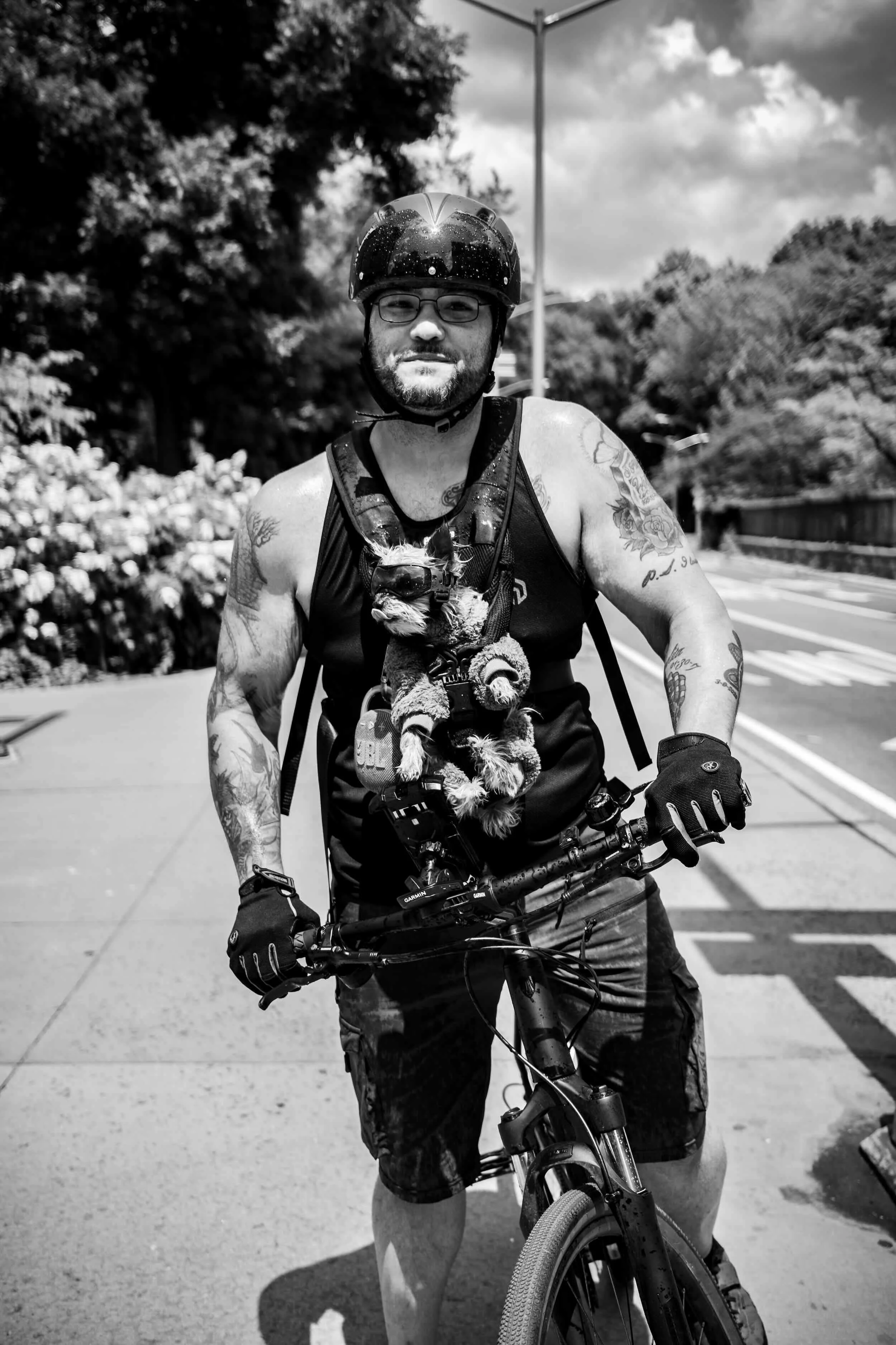 A man with glasses and tattoos riding a mountain bike outdoors wearing a helmet and sleeveless shirt, holding the bike’s handlebars, with trees and a cloudy sky in the background, captured in black and white.