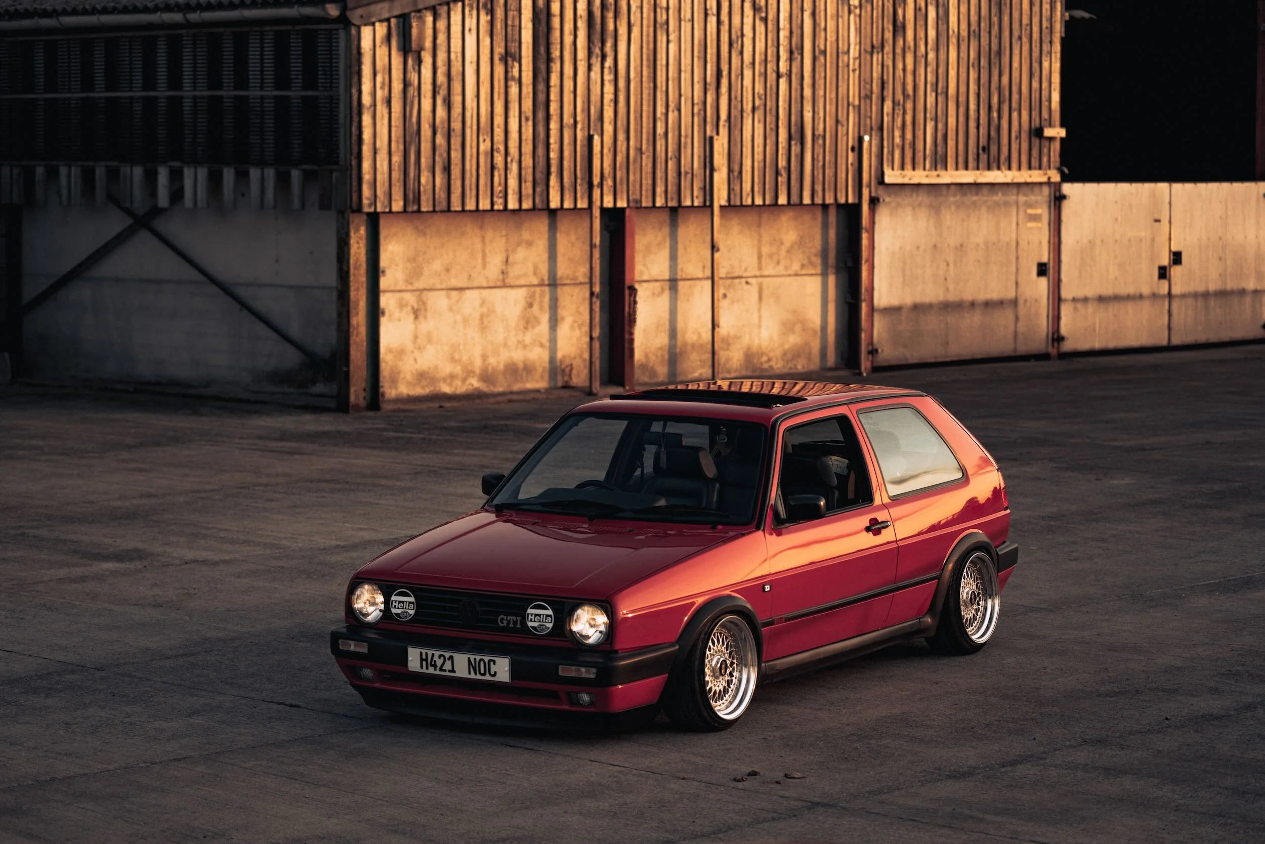 A red Volkswagen GTI parked in an empty lot with a wooden and concrete building in the background during sunset.