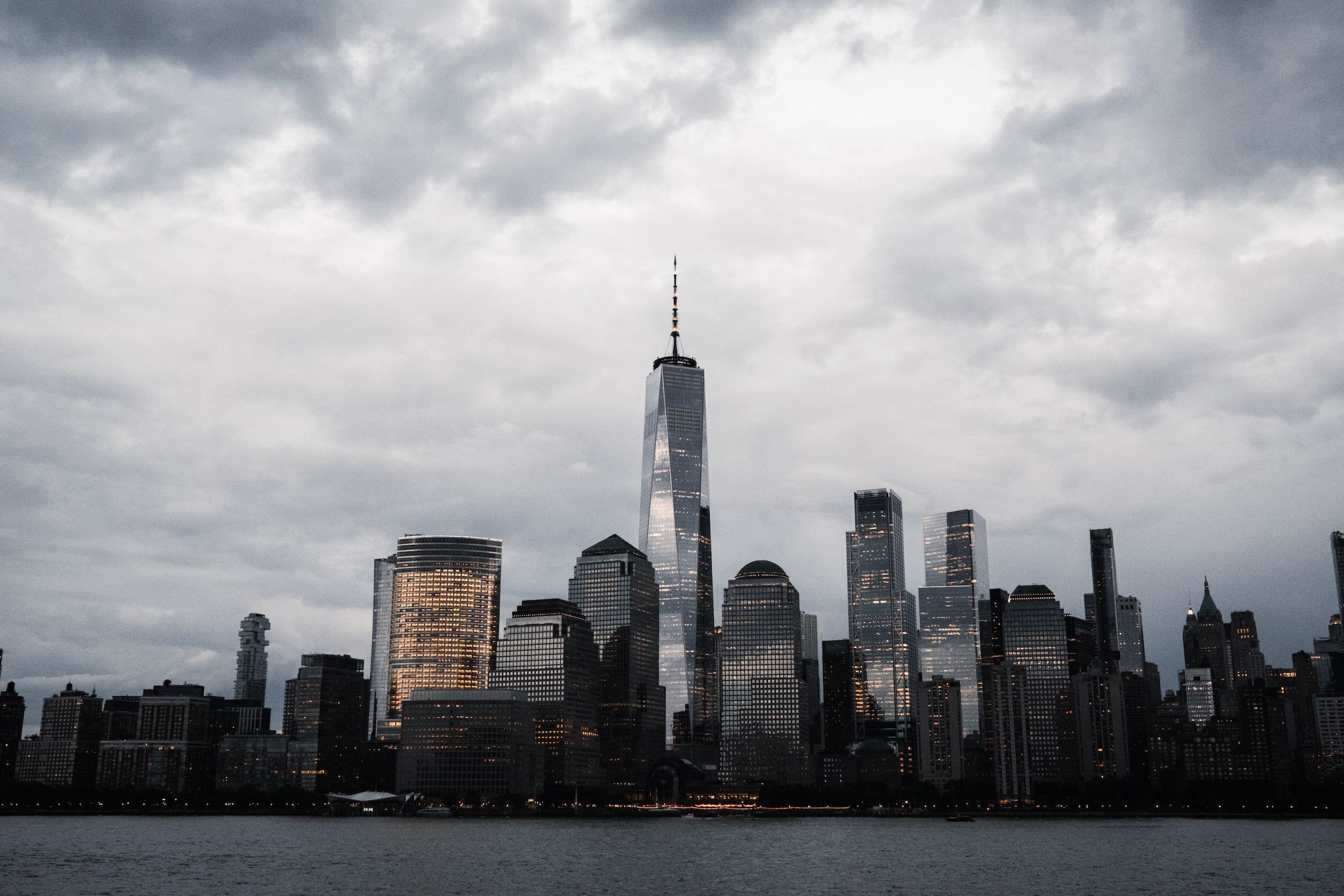 City skyline with tall skyscrapers, including the One World Trade Center, under a cloudy sky, seen from across a body of water.