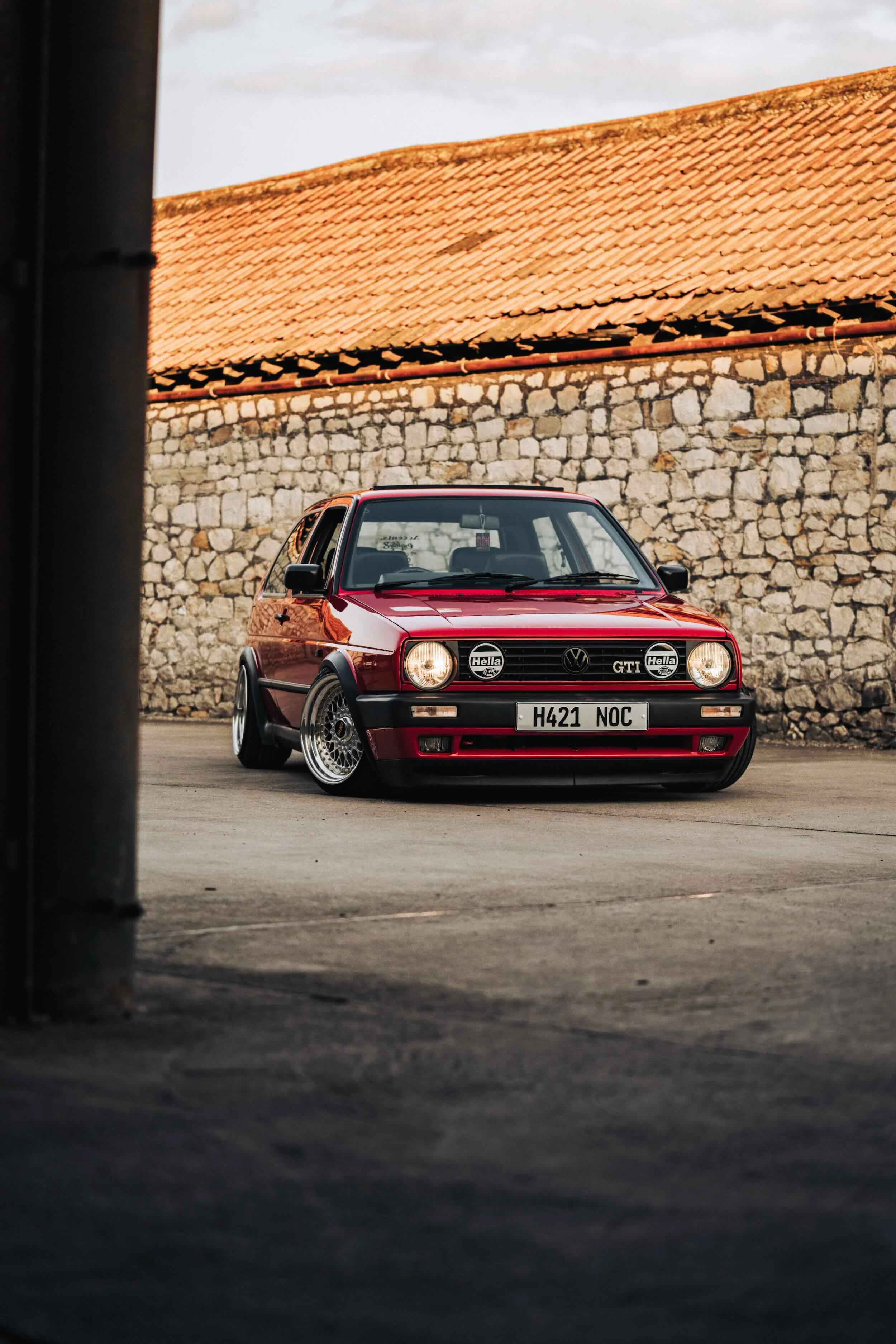 A red Volkswagen Golf GTI parked in front of a stone wall with a tiled roof, bathed in warm sunlight, with a stone wall and cloudy sky in the background.